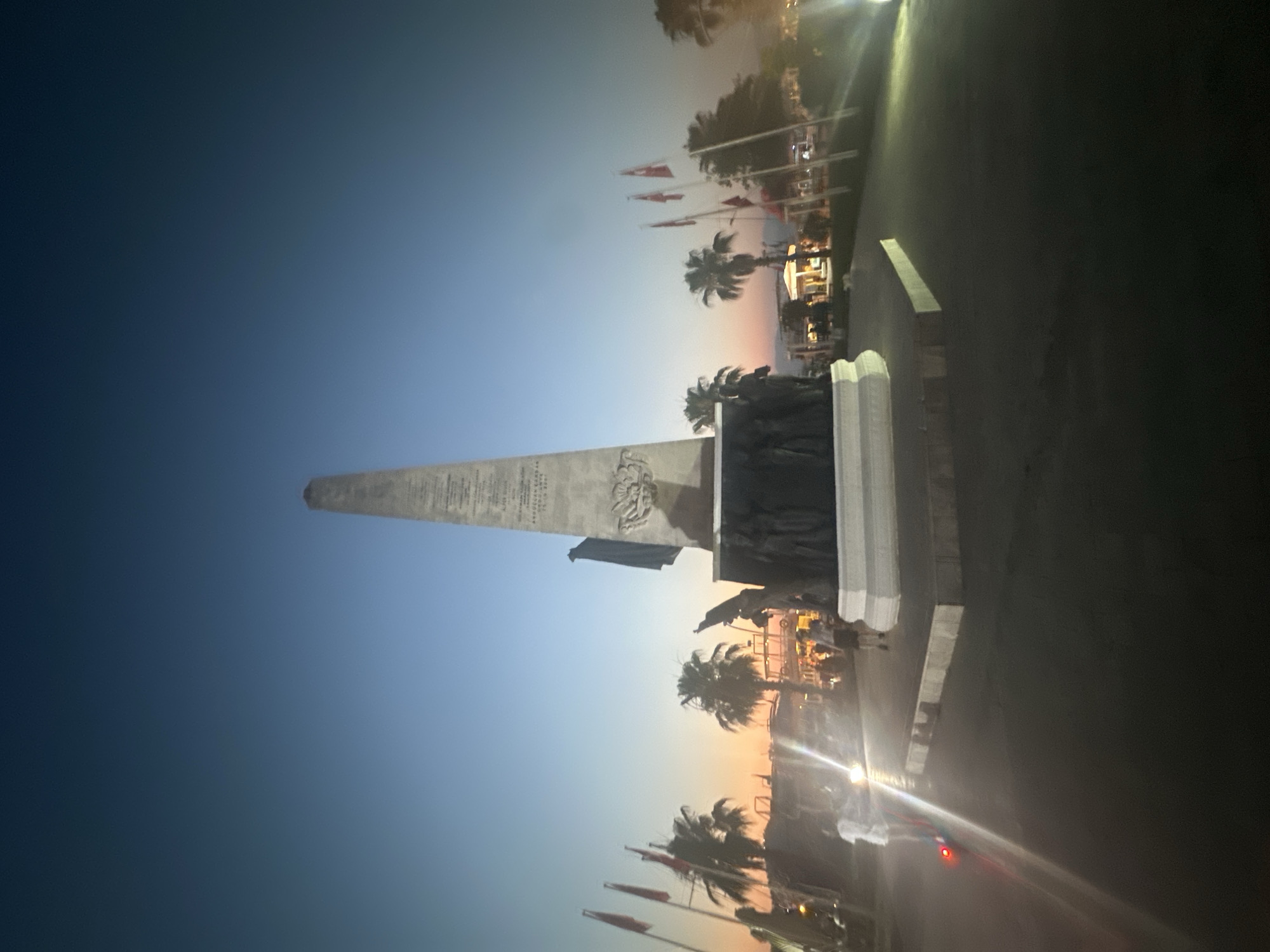 The image shows the Ataturk Monument in Fethiye, Turkey, at dusk. The monument is a tall obelisk with a statue of a figure at its base, surrounded by a group of bronze figures. Several Turkish flags are flying on poles to the left of the monument. Palm trees and some buildings are visible in the background. In the foreground, two people are walking, and another person is riding an electric scooter. The sky is a gradient of deep blue to orange, indicating the time of sunset or sunrise. The lighting suggests artificial lights are on in the background, illuminating the street and some of the buildings.