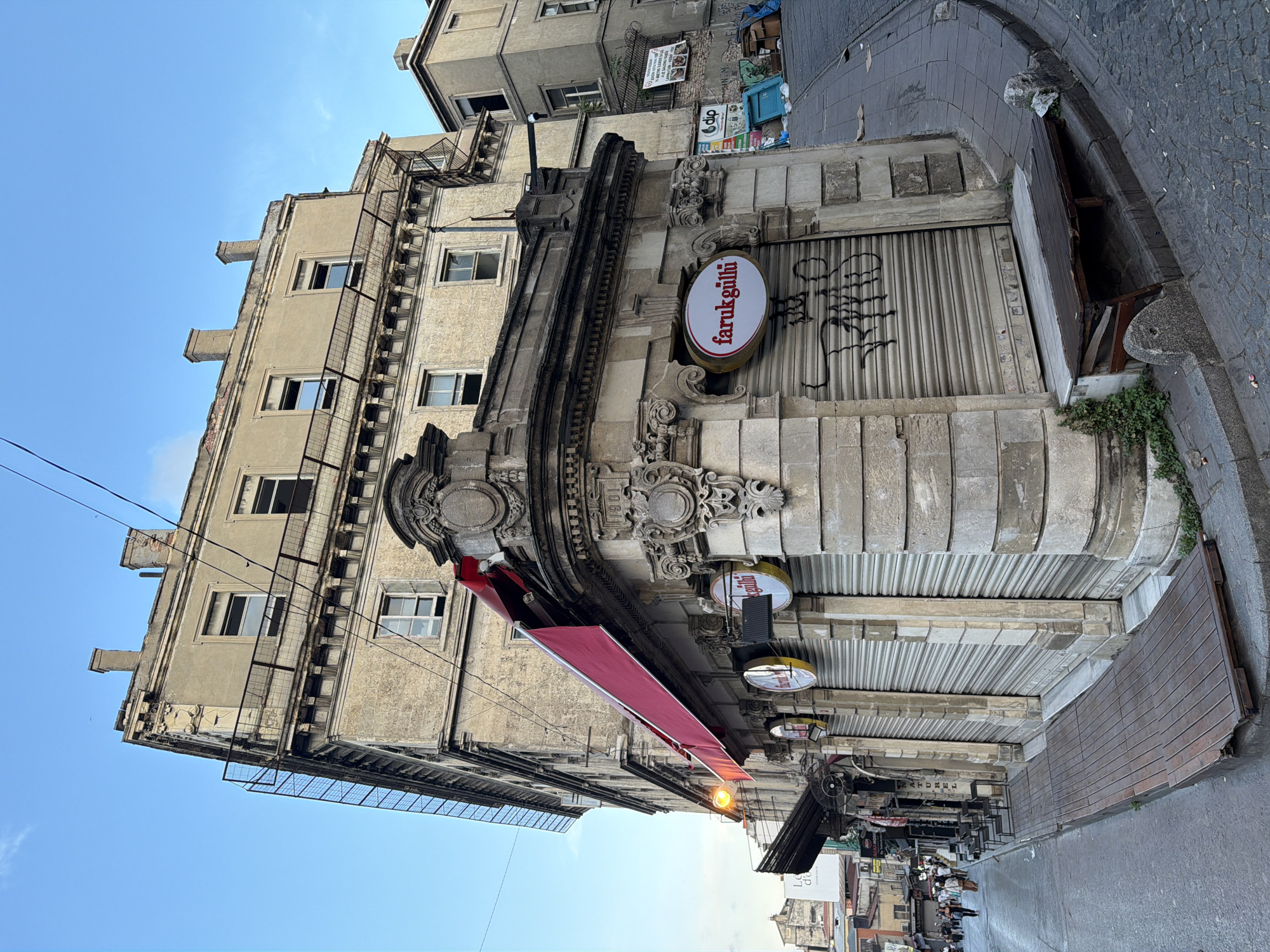 A slightly fisheye perspective captures an early evening street scene in Istanbul, Turkey. The focus is on a corner building with faded yellow stone facades and ornate architectural details, including carved cornices and decorative window surrounds. A bright red awning stretches across the storefronts, which are covered by closed, corrugated metal security shutters marked with graffiti. Two circular signs are visible; one reads "farukgüllü" in red lettering on a white background, and another is a yellow circle with black lettering. A simple wooden bench sits on the curb in front of the shuttered storefronts, showing signs of disrepair. To the right, the street slopes upwards, paved with cobblestones, and a second, taller building with similar architectural features and boarded windows stands behind the main subject. Further down the street, to the left, the scene opens to reveal more buildings, pedestrians walking, and some market stalls. Overhead, a clear blue sky with a few wispy clouds is crisscrossed by electrical wires. The overall impression is one of a historic, somewhat aged urban environment.