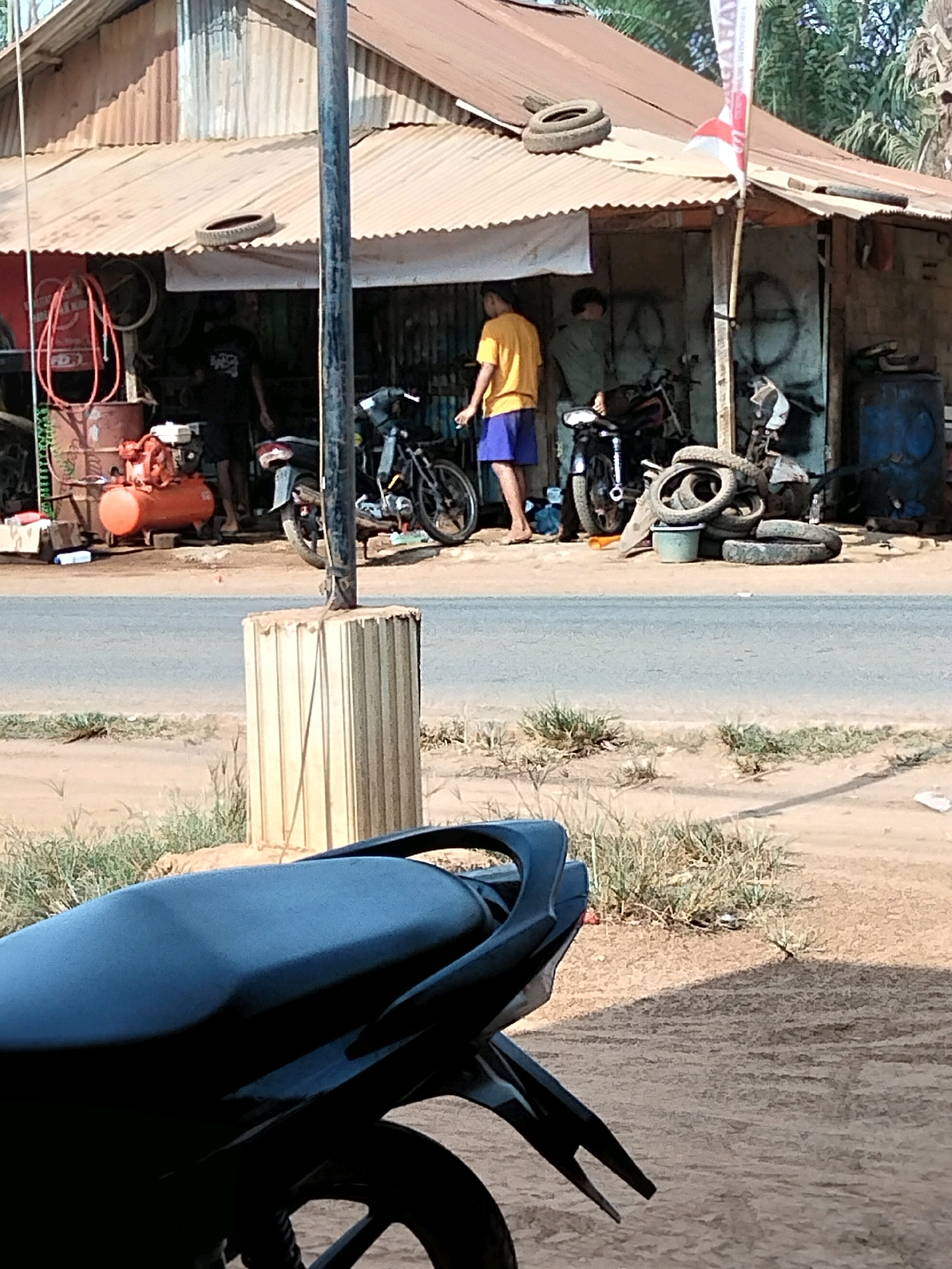 The image depicts an outdoor scene in Sidomulyo, Indonesia, featuring a roadside motorcycle repair shop. In the foreground, the black seat and tail of a motorcycle are visible. Behind it, a paved road separates the viewer from the shop. The shop itself is a modest structure with a corrugated metal roof and walls, appearing to be made of salvaged materials. Various items are present outside the shop, including an orange air compressor, a red hose coiled on a barrel, several used tires stacked and piled, and a blue barrel. Two men are standing near the entrance of the shop, one wearing a yellow shirt and blue shorts, and the other a green shirt. They are positioned near motorcycles, suggesting they are engaged in repair work. A red and white Indonesian flag is affixed to a pole near the shop. The surrounding environment is characterized by dirt ground and sparse vegetation.