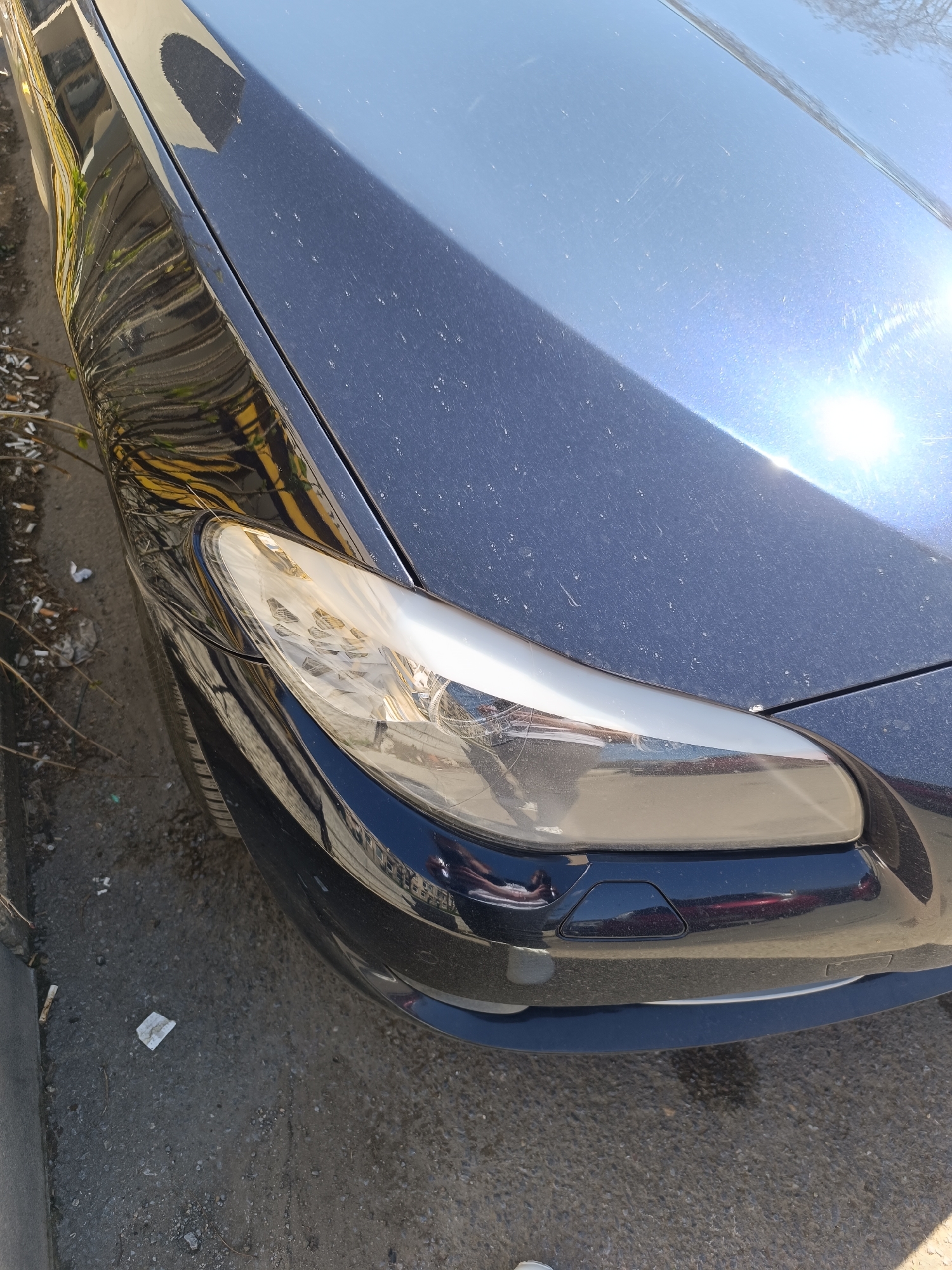 The image depicts the front of a dark blue car, possibly parked on a street in Sofia, Bulgaria. The car is dusty, with the front headlight prominently featured. The area beside the car appears to be littered with cigarette butts and other debris. The reflections on the car surface suggest an urban environment.