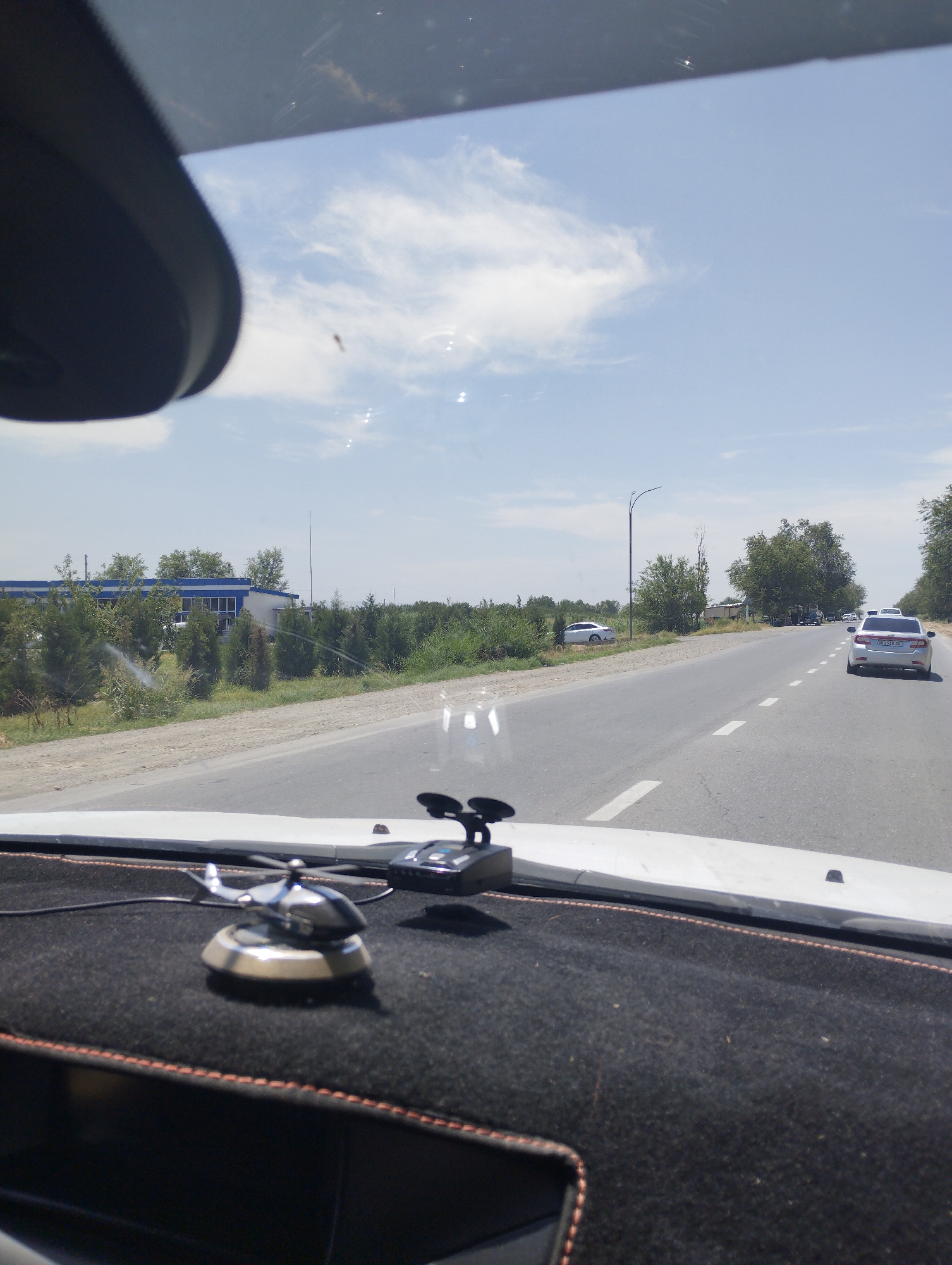 The image depicts a view from inside a vehicle traveling on a paved road. In the foreground, on the dashboard, there is a metallic helicopter figurine and a black electronic device, possibly a radar detector. The road extends into the distance, with a white dashed line marking the lanes. On the right side of the road, several cars are visible, including a silver sedan with a license plate. The left side of the road features a dirt shoulder, followed by a line of trees and a building with a blue roof and white trim. Beyond the trees, the sky is a clear blue with scattered white clouds. The overall scene suggests a sunny day in a semi-urban or rural area. This view is consistent with the area around Bustan, Uzbekistan, known for its roads lined with trees and low-lying buildings.