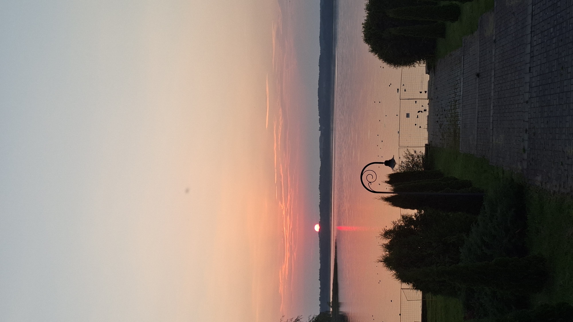 The image captures a serene sunset over a lake, likely Lake Myadzyel, in Myadzyel, Belarus. The sky exhibits a gradient of soft hues, transitioning from pale blue at the top to warm peach and pink near the horizon. Thin, wispy clouds are scattered across the sky. The sun, a vibrant red orb, is partially submerged below the horizon, casting a strong, shimmering reflection on the calm water. The water's surface is smooth, with subtle ripples and a warm, rosy glow reflecting the sky.
Along the lakeshore, a line of dark green coniferous trees and bushes forms a dense natural barrier. In the foreground, a paved pathway made of interlocking gray bricks leads down towards the lake. Flanking the pathway are neatly trimmed cypress-like trees. A decorative black lamppost with a curved arm and a vintage-style lamp is positioned to the right of the pathway, its silhouette stark against the twilight sky. A temporary metal fence runs along the edge of the property bordering the lake. The overall atmosphere is tranquil and picturesque, suggesting a peaceful evening setting.