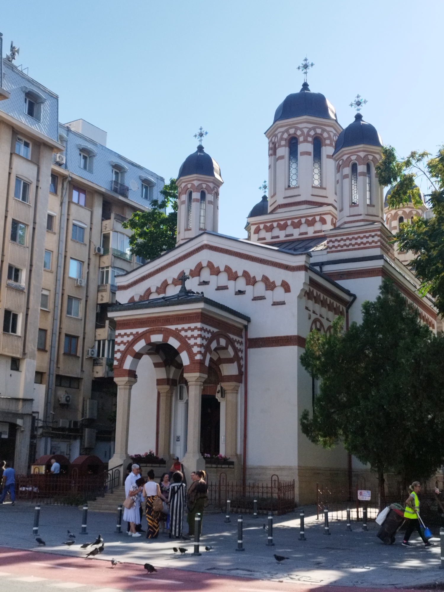 This is a medium shot, taken outdoors on a sunny day, of a white and red Orthodox church with multiple domes, typical of Bucharest, Romania. The church features arched windows and decorative brickwork. To the left of the church, a modern apartment building with balconies and grey tiled roofs is visible. In the foreground, several people are standing and talking on a paved area, with bollards and a red painted strip on the ground. Pigeons are scattered on the ground. To the right, a woman wearing a high-visibility yellow vest is pulling a large wheeled bin, holding a blue object, presumably a broom or cleaning tool. The scene appears to be a city street or square.