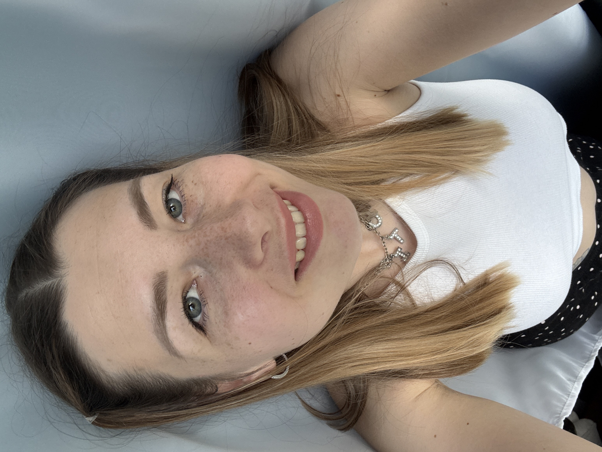 A young woman is depicted in a close-up selfie, lying down and looking upwards towards the camera with a bright smile. She has light brown hair, which is parted on the left side of her head and flows around her. Her eyes are light-colored, and she has visible eyeliner and well-groomed eyebrows. Numerous freckles are scattered across her nose and cheeks. She is wearing a white ribbed tank top and a necklace featuring large, silver, block letters that spell "LOYALTY." A small silver hoop earring is visible on her right ear. Part of a black garment with white polka dots can be seen on her right side, possibly a skirt or dress. The background is a plain, light blue-grey surface, suggesting an indoor setting. The overall mood is cheerful and relaxed. This image was taken in Moscow, Russia.