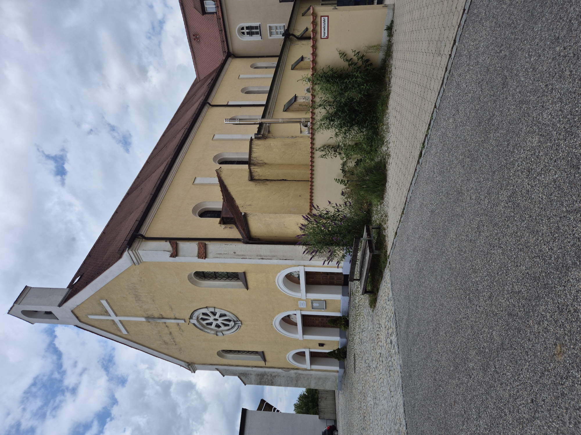 The image depicts the exterior of a yellow church building with a white cross on its facade, located in Landshut, Germany. The church has a bell tower with a white spire and a red tiled roof. The facade features arched doorways and windows, including a circular window with a rose design above the entrance. In front of the church, there is a cobblestone courtyard with a wooden bench. Lush green bushes and plants are visible along a low, red-tiled wall. To the left, a portion of a modern building is visible, along with a parked car. The sky is partially cloudy. A sign with the text "Feuerwehrauffahrt" (fire department access) is attached to the wall. The scene is an outdoor shot, taken from a low angle, with the church as the primary subject.
