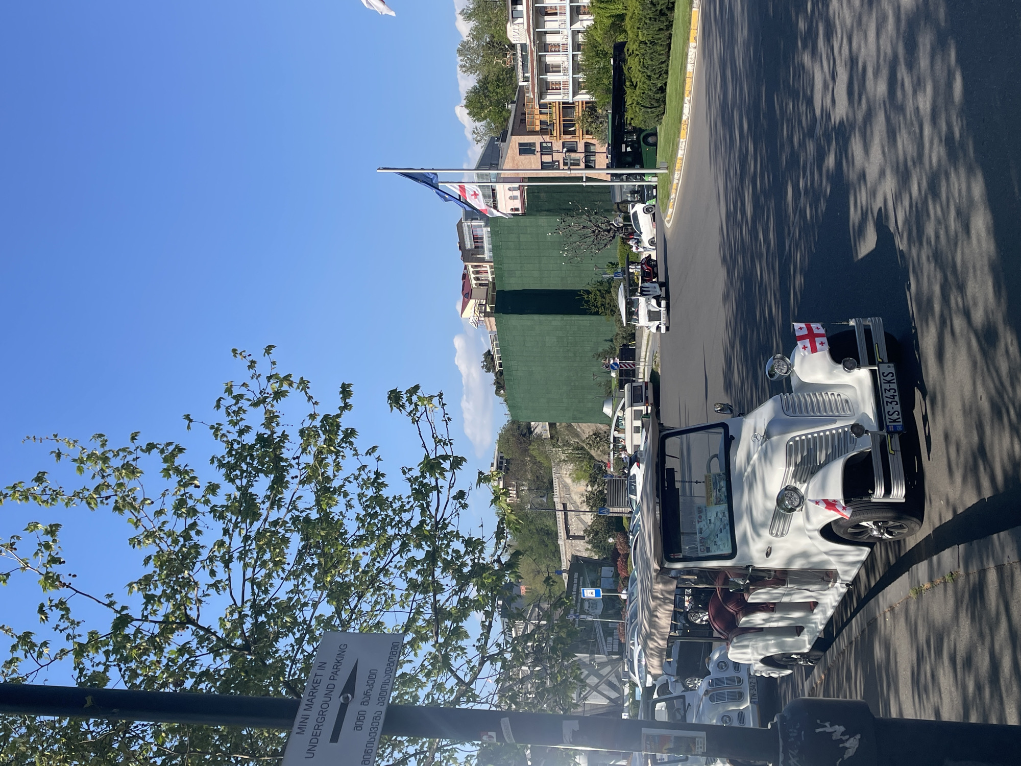 The image captures a bright, sunny day in Tbilisi, Georgia, showcasing a street lined with white vintage-style tour vehicles. The scene is oriented horizontally, with a clear blue sky above and strong sunlight creating distinct shadows of trees on the asphalt road.
In the foreground to the left, a tree with green leaves partially frames the view. Along the right side of the street, multiple open-top, white cars are parked. The closest vehicle prominently displays a Georgian flag on its front bumper and has the license plate "KS-343-KS". Another car's license plate, "TN-342-T", is also visible. Text suggesting "City Tour" can be partially seen on one of the vehicle's sides.
Further down the street, a flagpole holds two flags: the national flag of Georgia and another blue flag. Behind the vehicles and flags, a variety of buildings are visible, including structures with traditional balconies and modern elements, some featuring green roofs. Lush green trees and hedges line the street and buildings.
In the lower left, a black pole supports a white sign. The sign features text in both English and Georgian. It reads: "MINI MARKET IN UNDERGROUND PARKING" and its Georgian equivalent, "მინი მარკეტი მიწისქვეშა ავტოსადგომში". The image depicts a calm, typical daytime scene, likely in a tourist area of the city.
