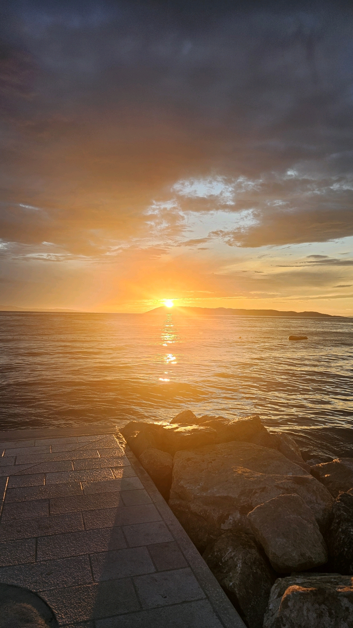 The image depicts a serene sunset over the Adriatic Sea, as viewed from a paved walkway and a rocky embankment. The sun, a brilliant orb, is low on the horizon, casting a warm golden glow that reflects vibrantly on the rippling water. Distant landmasses, likely islands or the mainland coast, are visible as silhouettes against the vibrant sky. The sky itself is a spectacle of color, transitioning from deep grays and blues at the top to fiery oranges and yellows near the horizon, with scattered clouds adding texture and depth. The foreground features a stone-tiled walkway on the left, leading towards a collection of large, rough-hewn rocks on the right, which form a natural barrier against the sea. This scene is likely located along the coastline of Općina Tučepi, Croatia, a region known for its picturesque Adriatic vistas.