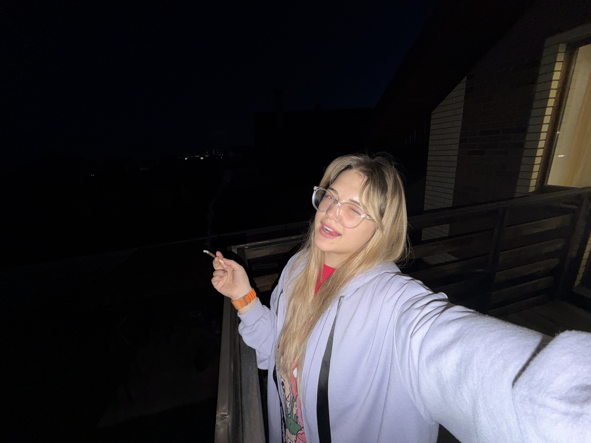 A young woman is captured in a selfie outdoors at night, likely on a balcony or deck. She has blonde hair and is wearing clear-framed eyeglasses, a light purple hooded sweatshirt over a red shirt, and an orange band on her left wrist. She holds a lit cigarette in her left hand, with a faint wisp of smoke rising from it. The woman is winking with her right eye and displaying a slight smile, conveying a playful and casual mood.
The background is dominated by darkness, indicative of nighttime. To the left, a dark expanse extends into the distance, with scattered, faint lights visible on the horizon, hinting at a settlement or structures in the area of Большая Шильна, Russia. To the right, a section of a brick building with a window is partially visible. A dark railing runs across the lower part of the frame, separating the woman from the shadowy surroundings. No discernible text is visible in the image.