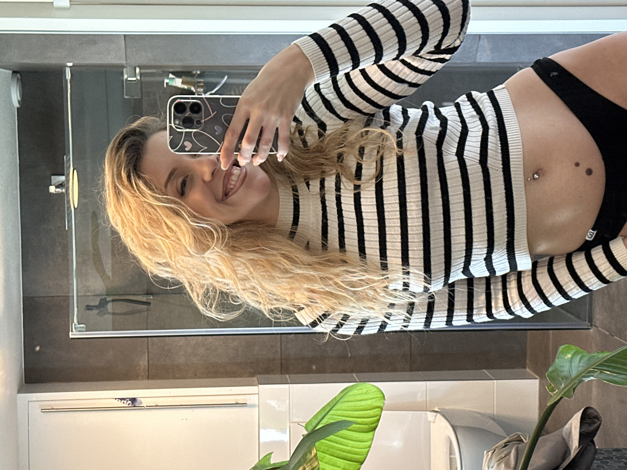A selfie taken in a bathroom. A blonde woman, wearing a black and white striped long-sleeved shirt and black underwear, is holding up a phone to take the picture. A bathroom sink, toilet, and houseplant can be seen in the background. The location appears to be a private residence. The image does not provide enough information to pinpoint the exact address or location within Dronten, Netherlands.