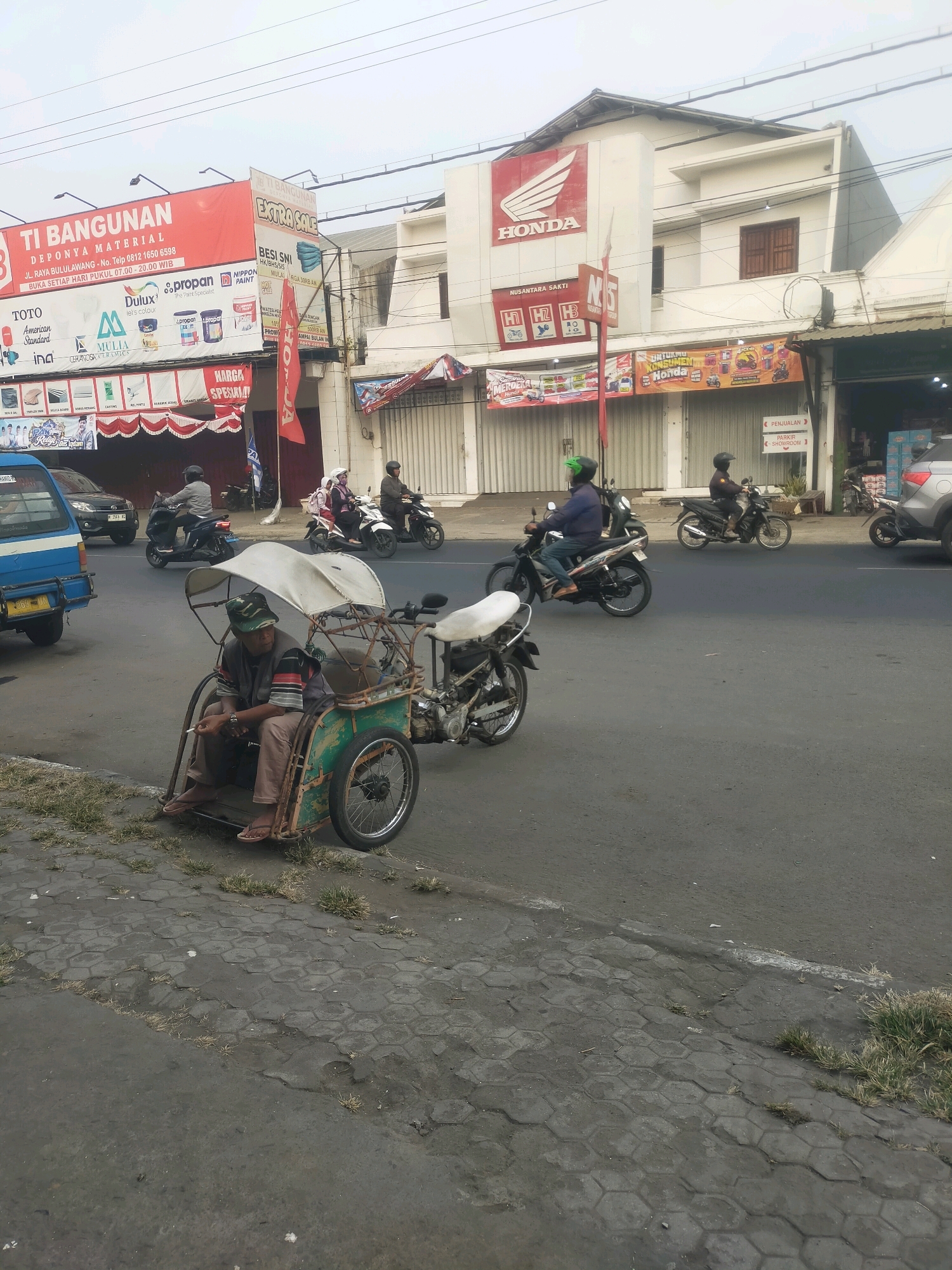 The image shows a street scene in Bululawang, Indonesia. In the foreground, a man sits in a pedicab, smoking a cigarette. The pedicab is green and has a white canopy. Behind the pedicab, several motorcycles are on the road. One motorcycle has a rider wearing a green helmet. Another motorcycle has a rider wearing a white headscarf and a pink backpack. Further back, there are buildings with signs. One sign says "TI BANGUNAN" and another has the Honda logo. There are also several other signs in Indonesian, indicating businesses and sales. The street is paved and there are some patches of grass growing through the pavement. The sky is overcast.