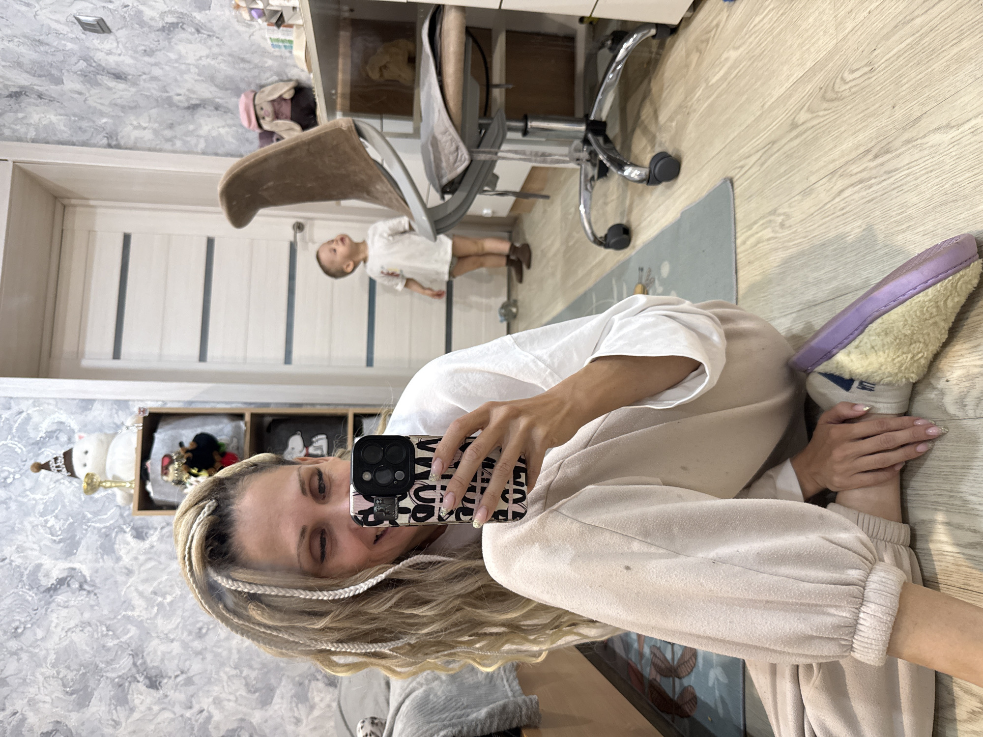 A woman is taking a selfie indoors in what appears to be a residence in Rusanovo, Russia. She is wearing a white top and beige sweatpants. Her hair is blonde with small white braids. She is holding a phone with a black and white patterned case. In the background, a young child wearing a white shirt and brown shoes stands near a desk and a chair. The room has light-colored wooden floors and white walls. There is a shelf with decorative objects visible. There is a mat on the floor. The overall lighting is bright. The image orientation is rotated 90 degrees.