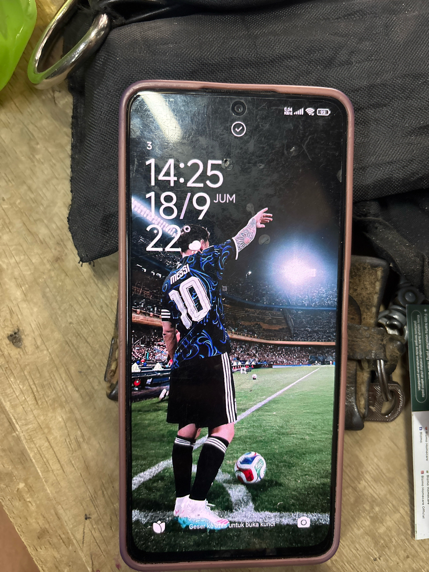 The image shows a smartphone displaying a lock screen with a soccer theme. The wallpaper features a soccer player, identified as "Messi" on his jersey, wearing a blue and black patterned shirt with the number "10" on the back. He is standing on a soccer field. The time displayed on the lock screen is 14:25, and the date is 18/9. The battery icon shows 93%. The phone is positioned on a wooden surface, with a dark fabric in the background. The scene appears to be indoors, possibly in a workshop or a similar environment, given the presence of tools and what looks like a lock. There are no people visible in the immediate vicinity of the phone, apart from the image displayed on the screen. The location is not identifiable as Kepolorejo, Indonesia, based solely on the visual information presented in the image.