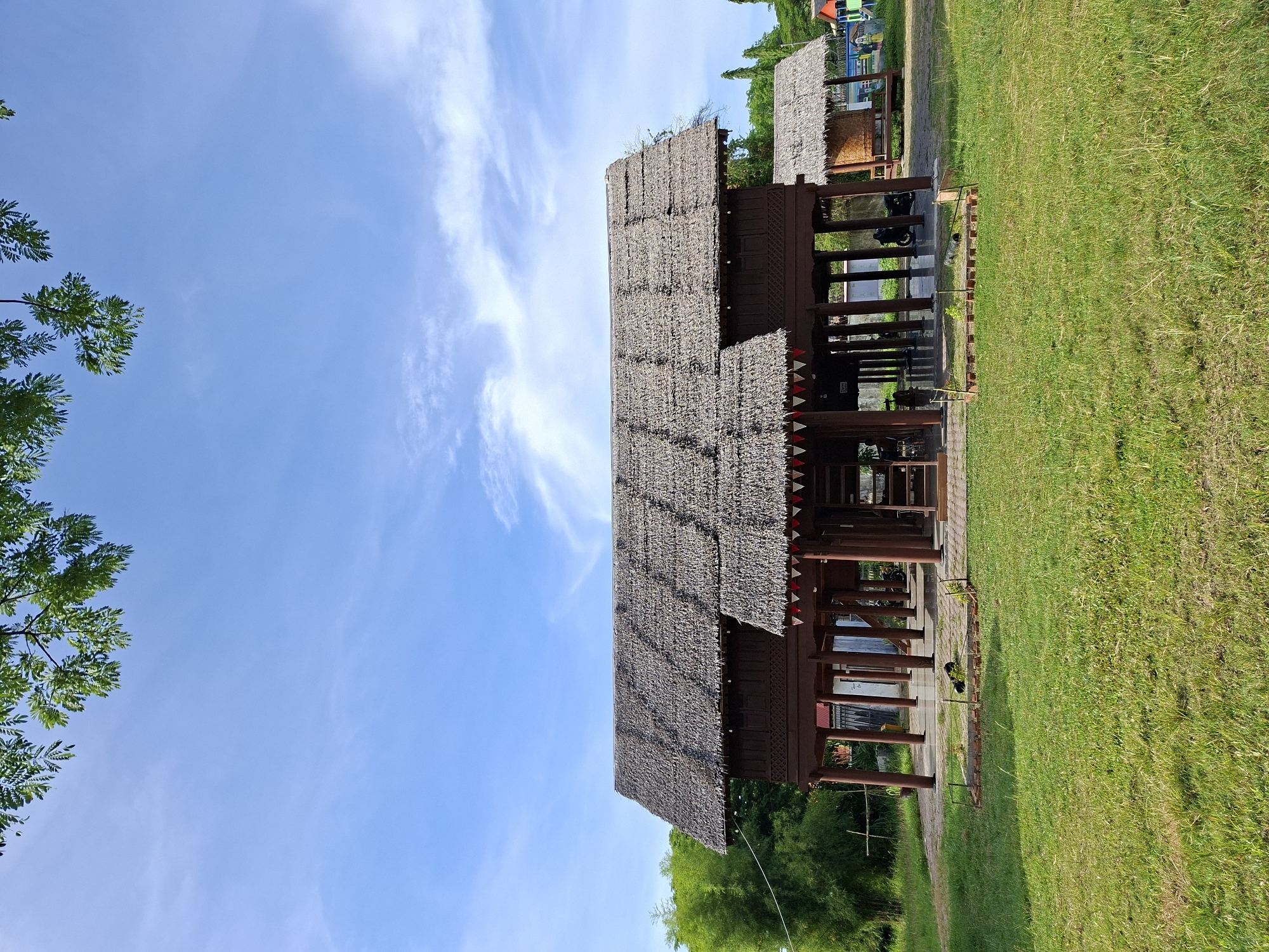 The image depicts a traditional Sumatran house in Lhokseumawe, Indonesia. The house has a thatched roof and is elevated on wooden pillars. It is surrounded by a green lawn and trees. The sky is blue with scattered clouds. There are a couple of other similar houses in the background.