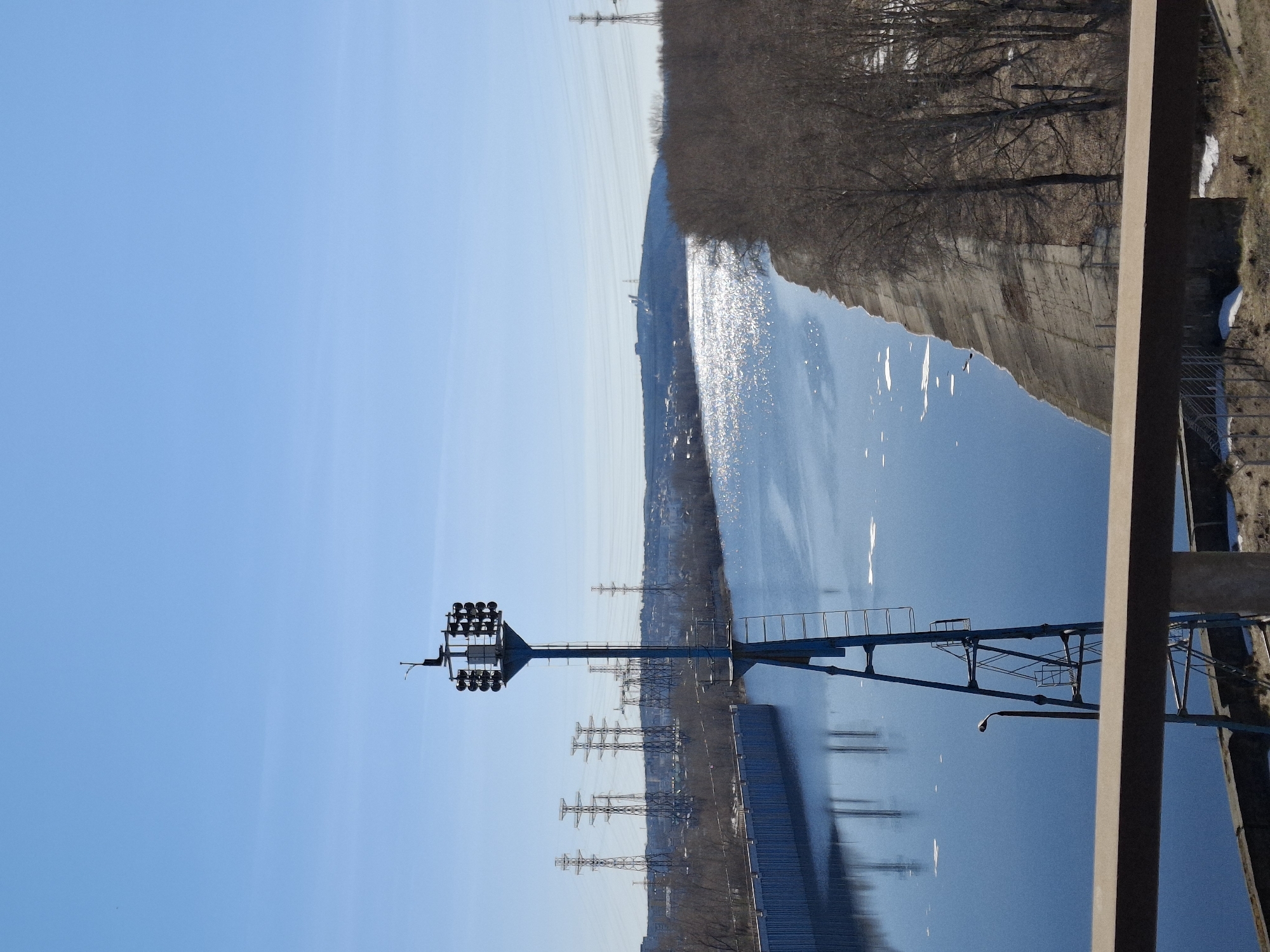 This media file captures an outdoor scene featuring a wide body of water, likely a river or large canal, under a clear, bright blue sky. The image is taken from an elevated perspective, possibly from a bridge or an embankment.
In the foreground, on the left side, a tall, dark-colored metal floodlight tower stands prominently, equipped with multiple clusters of lights at its top. On the far right, a large, light-brown concrete beam and some metal railings, part of a larger structure, are visible, partially framing the scene.
The water surface reflects the bright sky and shows numerous glinting points where sunlight hits, indicating a sunny daytime. Along the distant shoreline, a line of bare trees, possibly signaling late autumn, winter, or early spring, stretches across the frame. Beneath these trees, several large electricity pylons are dotted along the bank, suggesting industrial or utility infrastructure. Faint outlines of distant buildings or structures are also discernible on the horizon. On the immediate right bank, patches of snow are visible on the brown, grassy ground, reinforcing the impression of a cold season.
No people or specific activities are depicted in the image. The scene conveys a sense of calm, with a mix of natural elements and human-made infrastructure. There is no visible text.