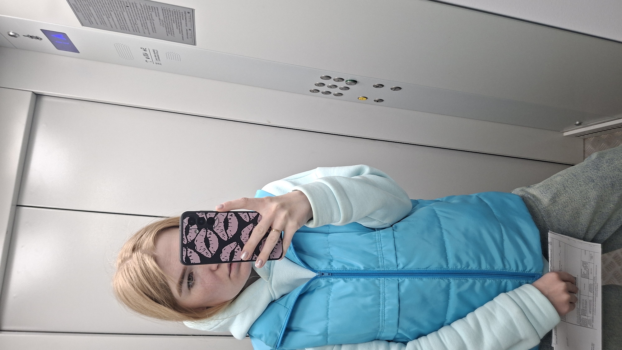A blond woman takes a selfie in an elevator in Chaikovsky, Russia. She is wearing a light blue hoodie under a blue puffer vest and grey sweatpants. Her phone has a black case with pink lips on it. The elevator is white with a panel of floor buttons and a call button. There is a notice in Russian above the buttons. The floor of the elevator is metal. She is holding some documents in her other hand.