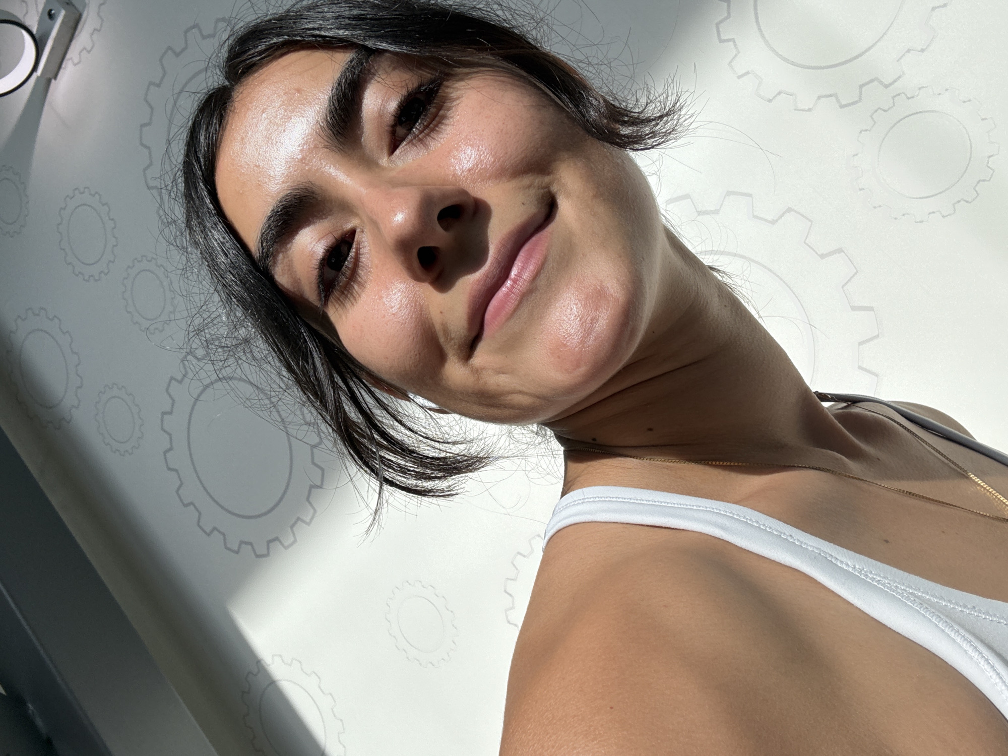 A close-up, tilted upward shot shows a woman with dark hair and a white tank top. She is smiling slightly. The background is a white wall with a repeating gear pattern. The environment suggests an indoor space, possibly in Puebla, Mexico, based on the light and architectural style. The lighting is bright, with shadows indicating a nearby light source. A gold necklace is visible on her neck.