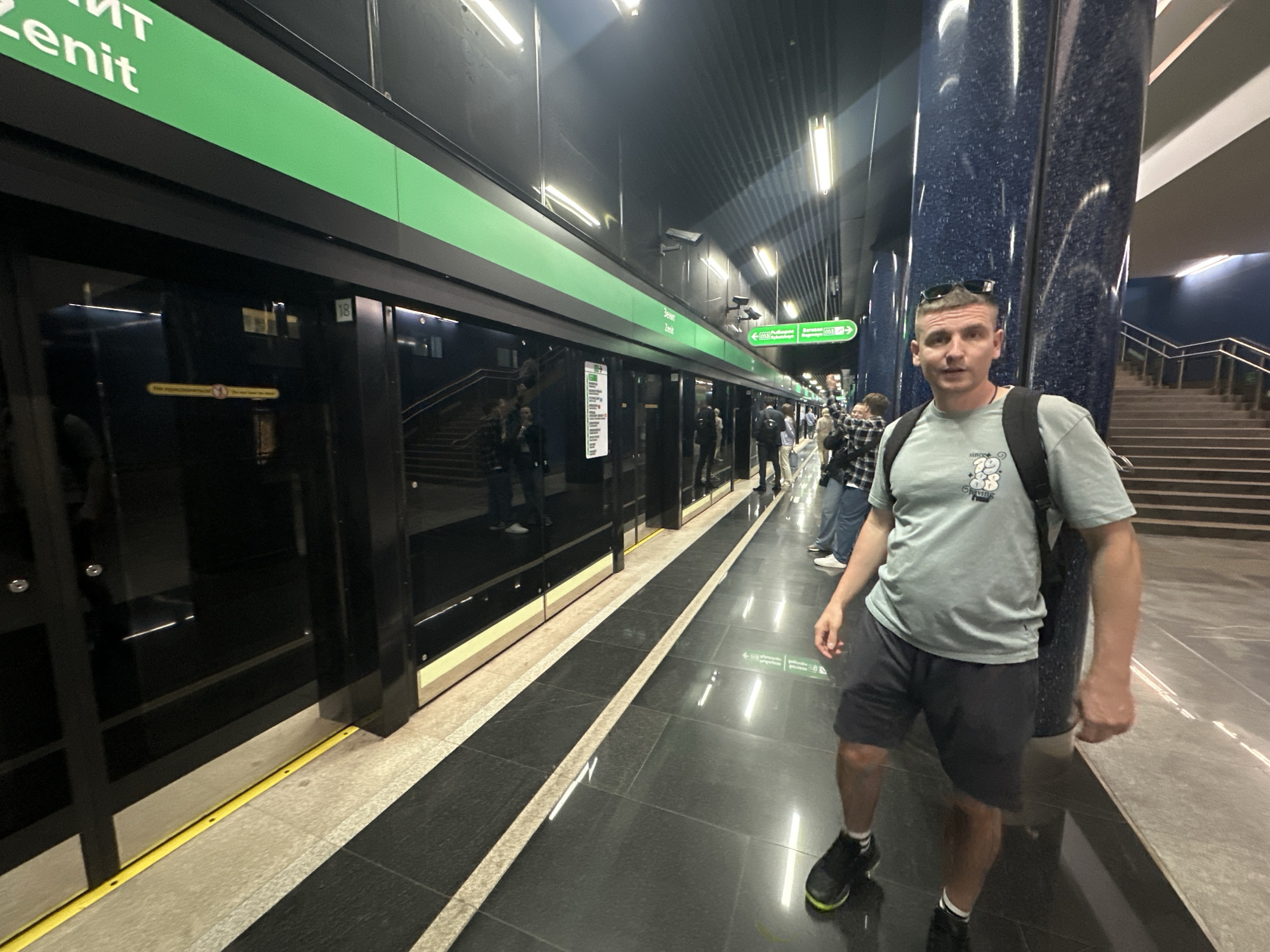 The image shows the platform of the Zenit metro station in Saint Petersburg, Russia. The station is modern, with platform screen doors that separate the platform from the tracks. A sign above the doors reads "Зенит Zenit" in white letters on a green background. The station is well-lit. The flooring is glossy and patterned with light and dark rectangles.
A man stands on the platform facing the camera. He wears a light green t-shirt, dark shorts, and dark sneakers with white socks. He has a backpack and sunglasses perched on his head. Other people are present on the platform, some near the stairs leading up from the platform. A blue pillar stands nearby, and stairs are visible leading up from the platform area.
