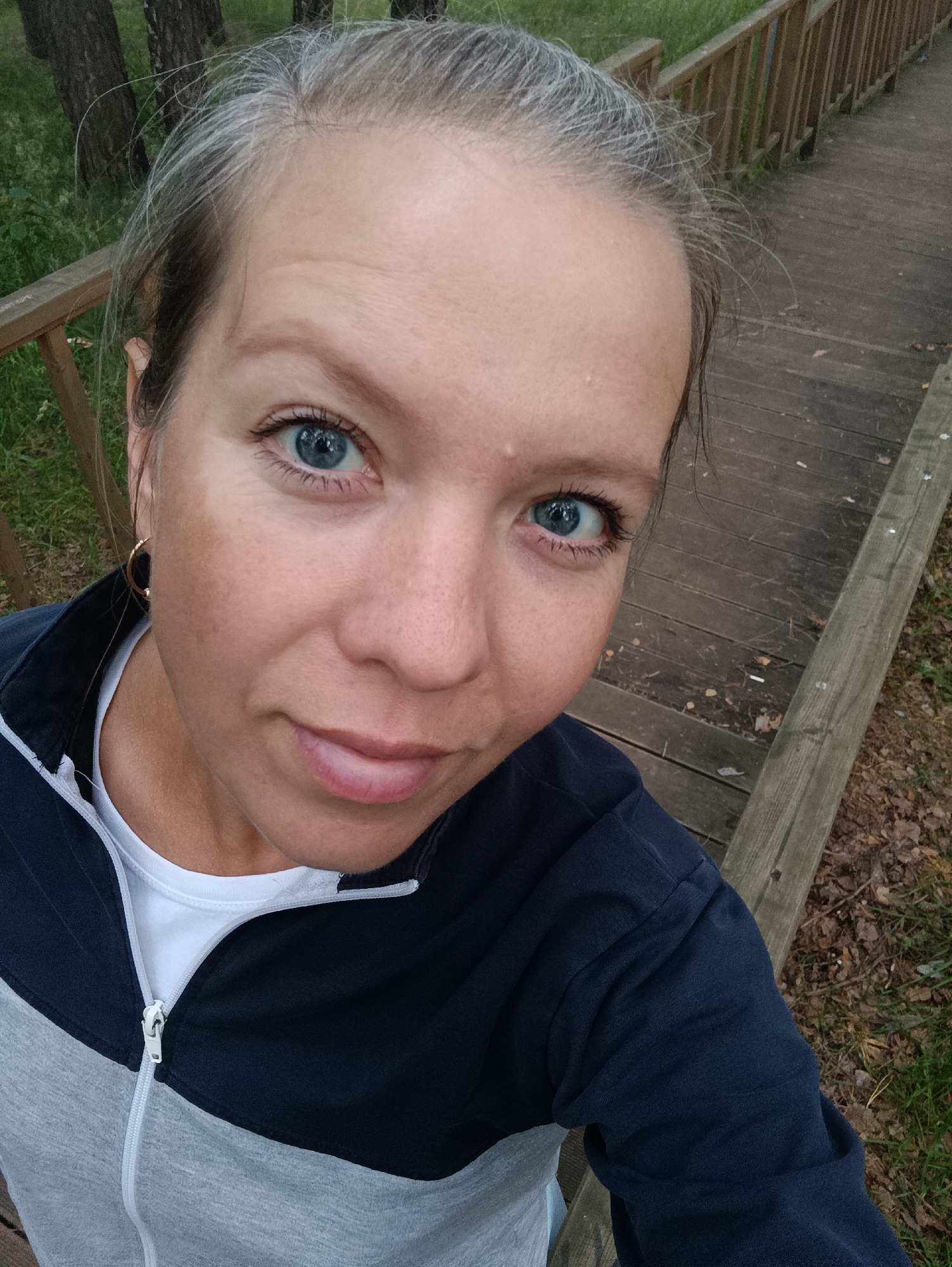 A medium shot captures a woman's face from the chest up as she takes a selfie outdoors. Her head is tilted slightly to her right, and her gaze is directed towards the camera. She has light skin with freckles, and her blue eyes are open. Her lips are slightly parted. She has gray hair pulled back from her face. She is wearing a dark blue and gray zippered jacket over a white t-shirt, and a gold hoop earring is visible in her left ear.
The background consists of a wooden boardwalk, with trees and foliage visible to the left and behind. The boardwalk appears to be in a natural setting, possibly a park or recreational area, and suggests a location in the city of Chaikovsky, Russia, given the context. The lighting indicates it is daytime.