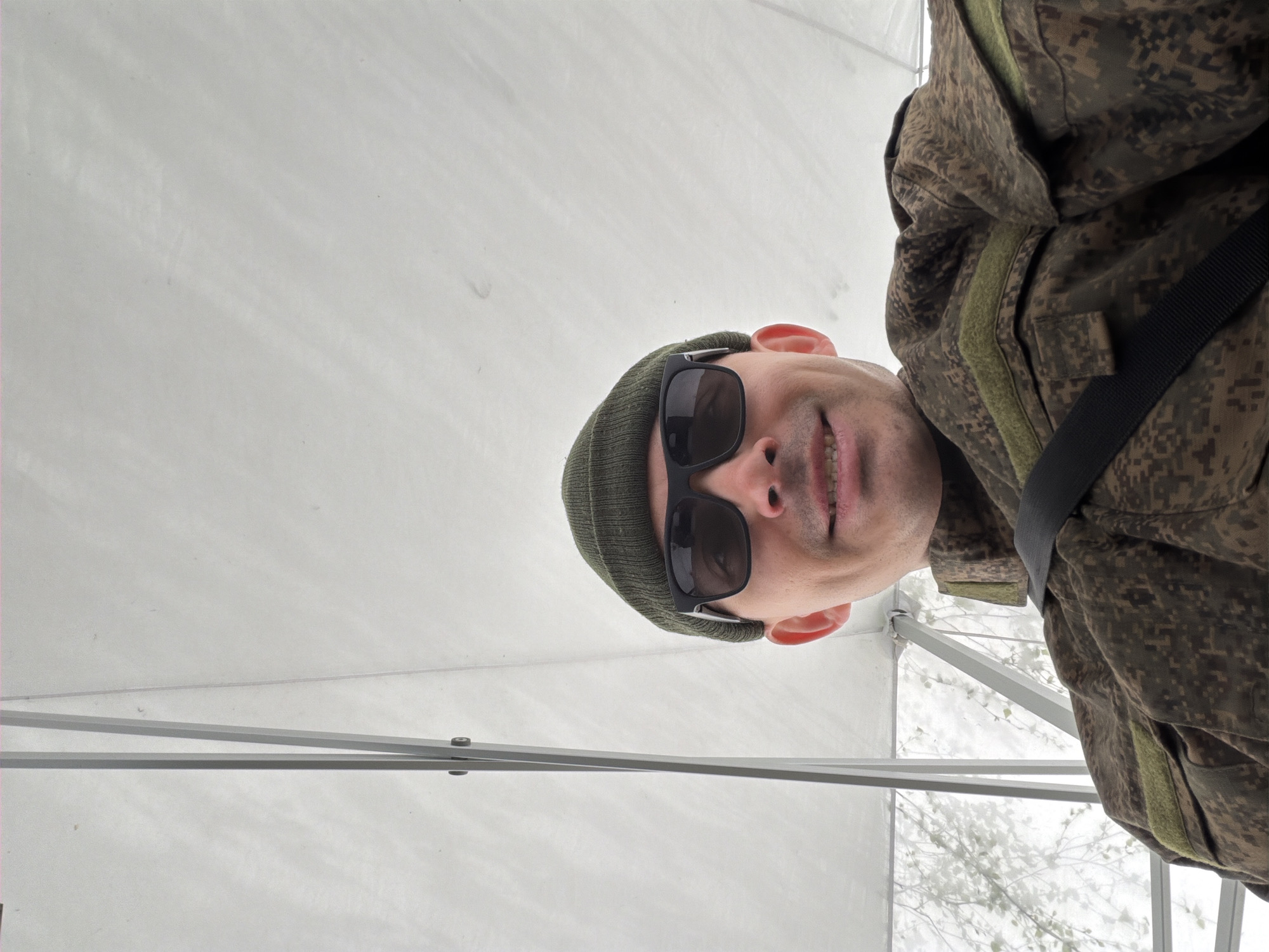 A low-angle shot captures a man in military fatigues, sunglasses, and a beanie, inside a white tent or structure. The location appears to be Izhevsk, Russia, evidenced by a possible Russian uniform. The man has light skin and is smiling. The background reveals a snowy scene with trees visible outside the tent. A black strap crosses his torso. Tent supports are visible in the foreground.