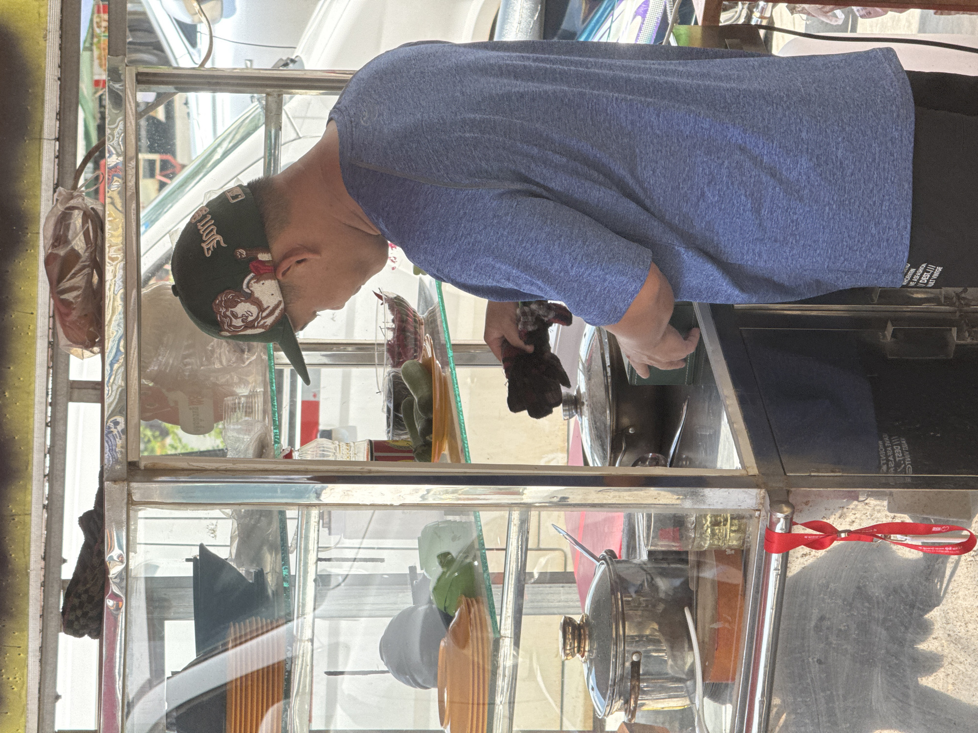 A person wearing a blue t-shirt and a dark green baseball cap is standing behind a food cart in Bavet Quarter, Cambodia. The food cart is made of metal and glass, with shelves displaying various items. On the top shelf, there is a brown bag of food. The second shelf holds orange plates stacked, a black bowl, and some green items. The third shelf contains a red bowl with small red items, and some green items on a plate. On the counter area of the cart, there are two metal pots with lids, a glass of yellow liquid, and a small metal tray. The person is holding a dark cloth in their right hand and appears to be interacting with the food on the counter. A red lanyard with some printing is hanging from the side of the cart. The background shows a blurred view of a street with vehicles.