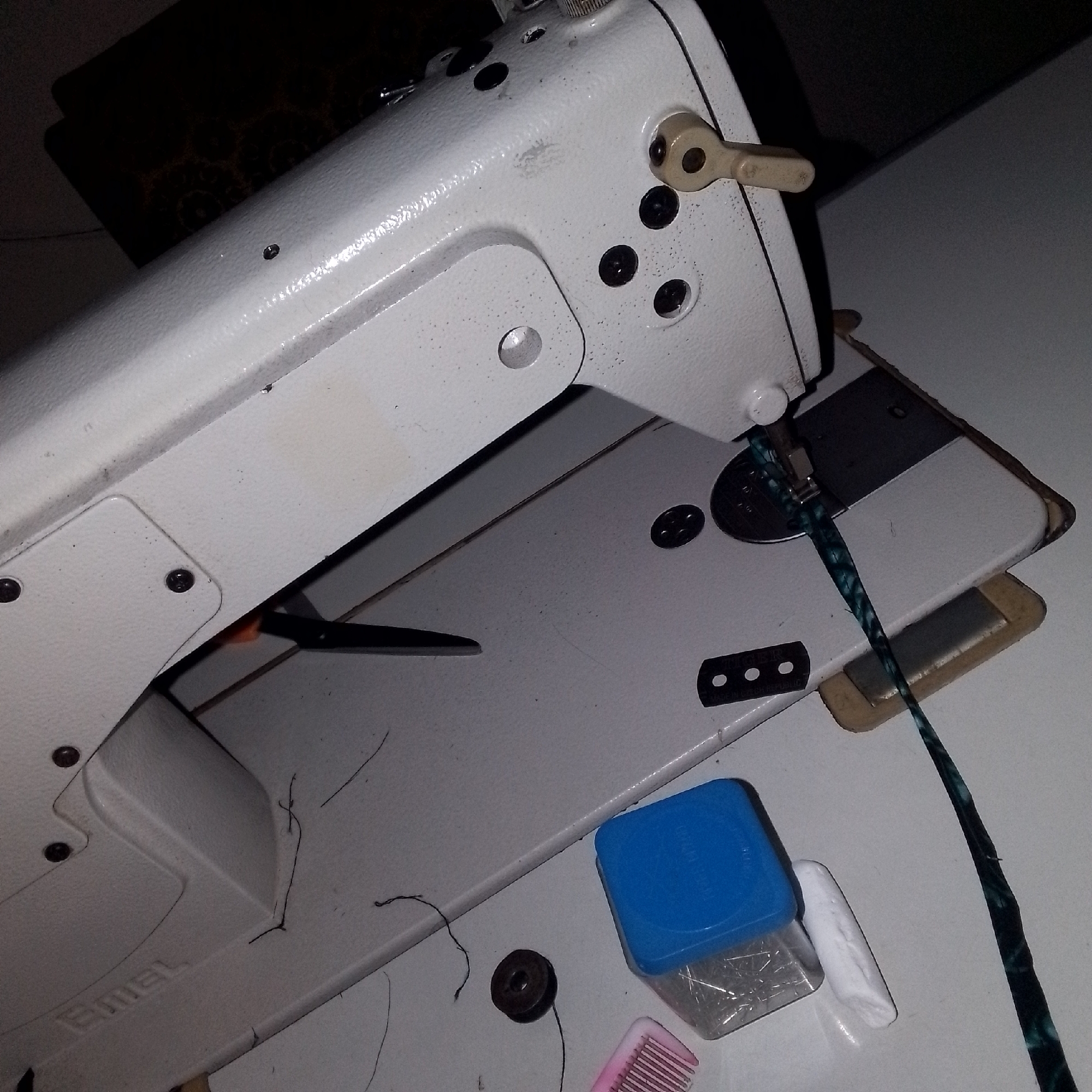 The image shows a close-up of a white industrial sewing machine. A dark, patterned cord or strap is visible, threaded through the machine's needle mechanism and hanging down. To the right of the sewing machine, on a white surface, are a blue plastic container filled with sewing pins, a white chalk-like object, and a pink object that appears to be a small comb or brush with teeth. A dark spool of thread with a strand of thread attached is also visible on the white surface. The environment appears to be an indoor space, possibly a workshop or a home where sewing is done, and there are some stray threads scattered on the surface. The background is dark and out of focus, suggesting a dimly lit area. There is no indication of Jauro Yinu, Nigeria, or any specific city.