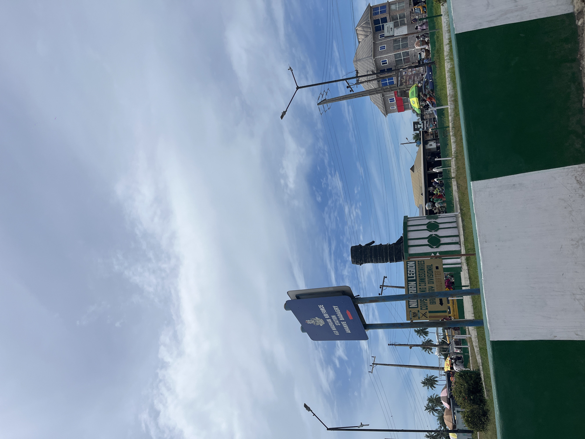 The image captures a scene in Badagry, Nigeria. The sky is partially covered by clouds. In the foreground, there is a sign indicating "653 NIGERIAN AIR FORCE STATION, AHANVE BADAGRY." Beside it is a sign for the "NIGERIAN LEGION CORPS," possibly related to military or veteran affairs. The environment includes utility poles and palm trees along the road. People, vehicles, and buildings can be seen in the background, suggesting a bustling street scene. A structure painted in green and white stands adjacent to the road.