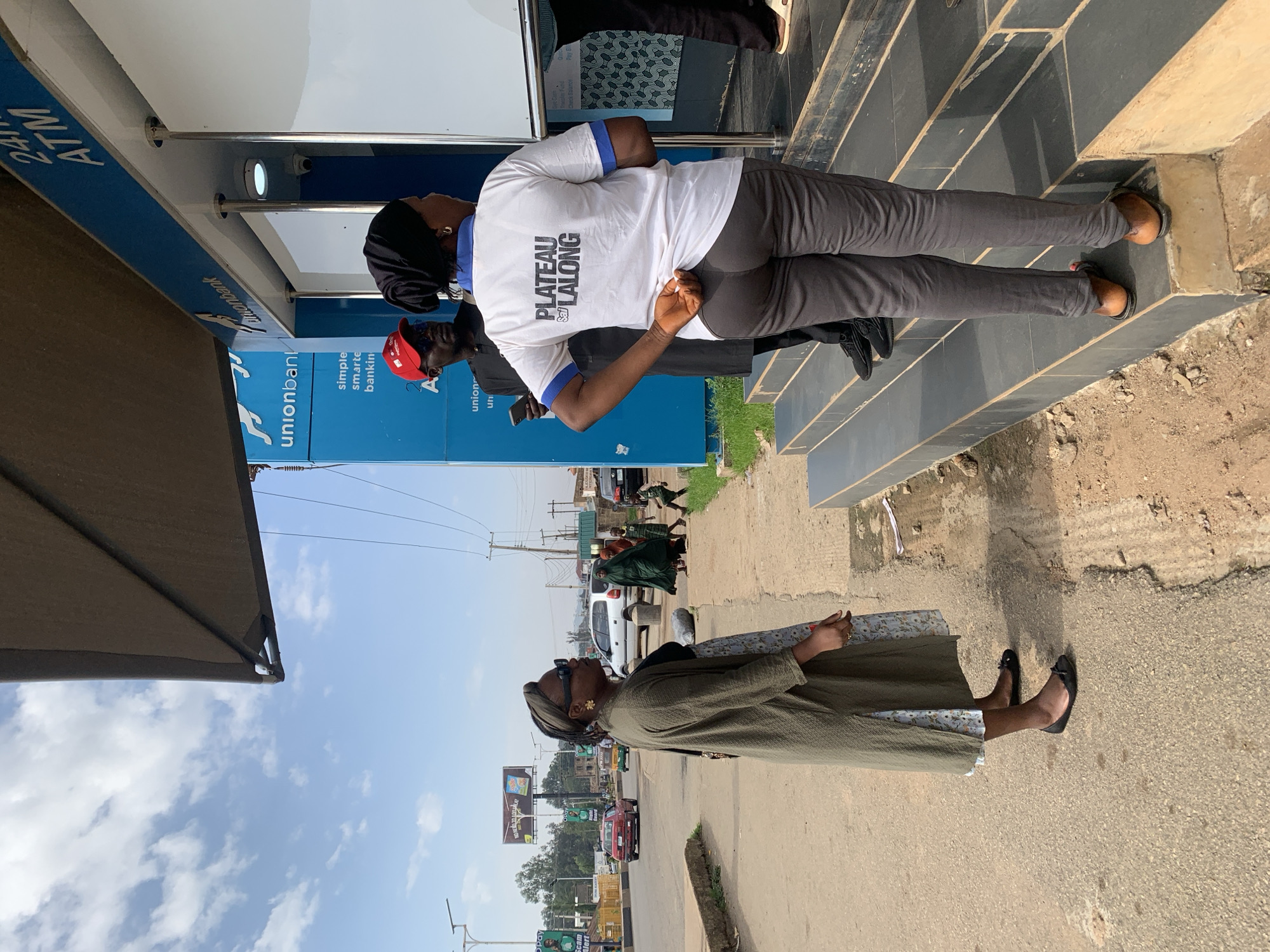 The image depicts a street scene in Jos, Nigeria, with a focus on individuals standing near a Union Bank ATM. In the foreground, a woman in a long, olive-green cardigan and a floral-patterned dress, wearing black ballet flats, stands on a paved street, looking towards the right. Behind her, a man in a red cap and sunglasses, wearing a dark shirt, is looking at his phone. To the right, a woman in a white t-shirt with "PLATEAU LALONG" printed on the back and dark grey pants stands on a set of steps leading up to the ATM. She is wearing brown sandals. Another person is partially visible in the background, wearing dark pants and black shoes. The Union Bank building features blue signage with "24H ATM" prominently displayed. In the middle ground, traffic, including cars and a tricycle, moves along a road. Further in the background, various buildings and structures are visible, along with utility poles and wires, under a partly cloudy sky. Several other individuals, some in traditional attire, are seen walking on the sidewalk in the middle ground. The overall environment suggests an urban setting with everyday street activity.