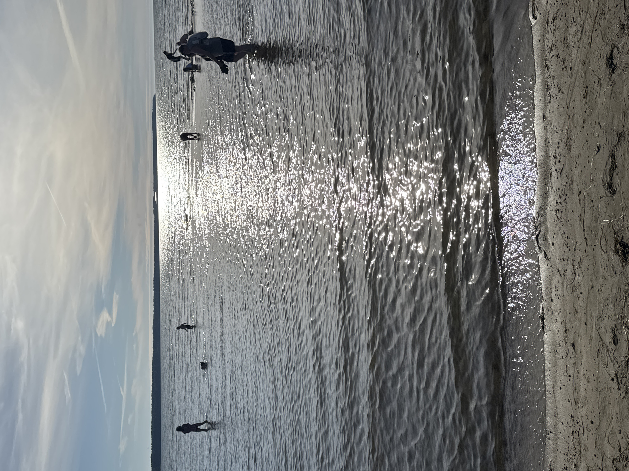 A group of people are wading in a body of water, possibly a lake or sea, near a sandy shore. The sun is bright, reflecting off the water's surface and creating a shimmering effect. The sky is partly cloudy, with some thin, wispy clouds. In the foreground, a person is standing in shallow water, with the lower half of their body submerged. Further out, other figures can be seen wading in the water, some appearing to be children or smaller individuals. One person appears to be near the shore, partially obscured by the water. The overall scene suggests a leisurely day spent by the water, possibly during the summer. The lighting indicates it is likely daytime, with the sun high enough in the sky to cause significant glare. The sandy shore, with some debris and dried vegetation, suggests a natural coastline. There is no visible text in the image.