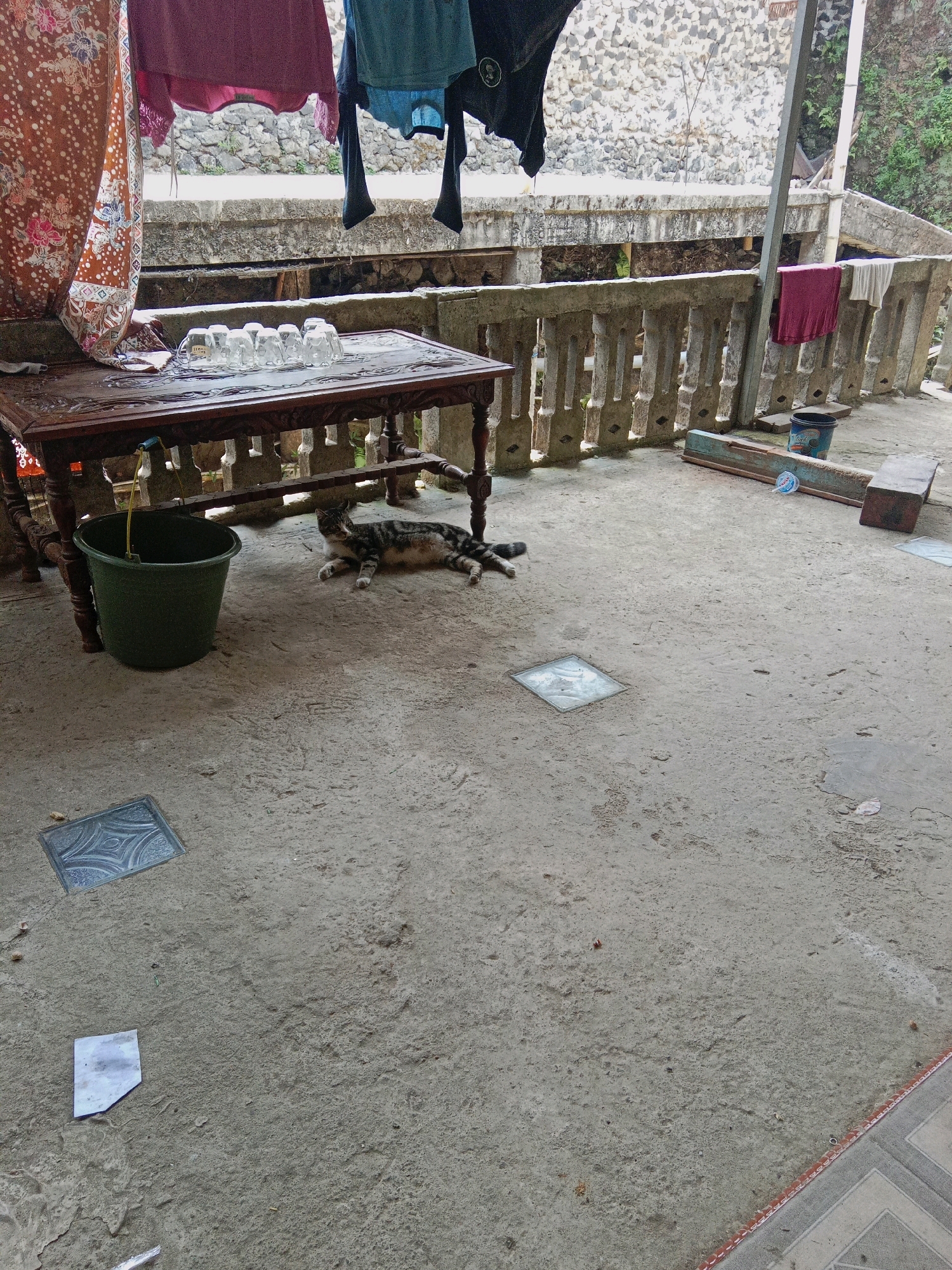 In Pulosaren, Indonesia, an outdoor scene depicts laundry hanging from a clothesline above a wooden table. On the table, several clear glasses are arranged. Beneath the table, a green bucket sits on the concrete ground, and a tabby cat is lying down. The background features a stone wall and a concrete railing. The ground is a mixture of concrete and dirt, with some square decorative tiles embedded in it. Various objects, including a wooden plank, a bucket, and a plastic bottle, are scattered around the area.