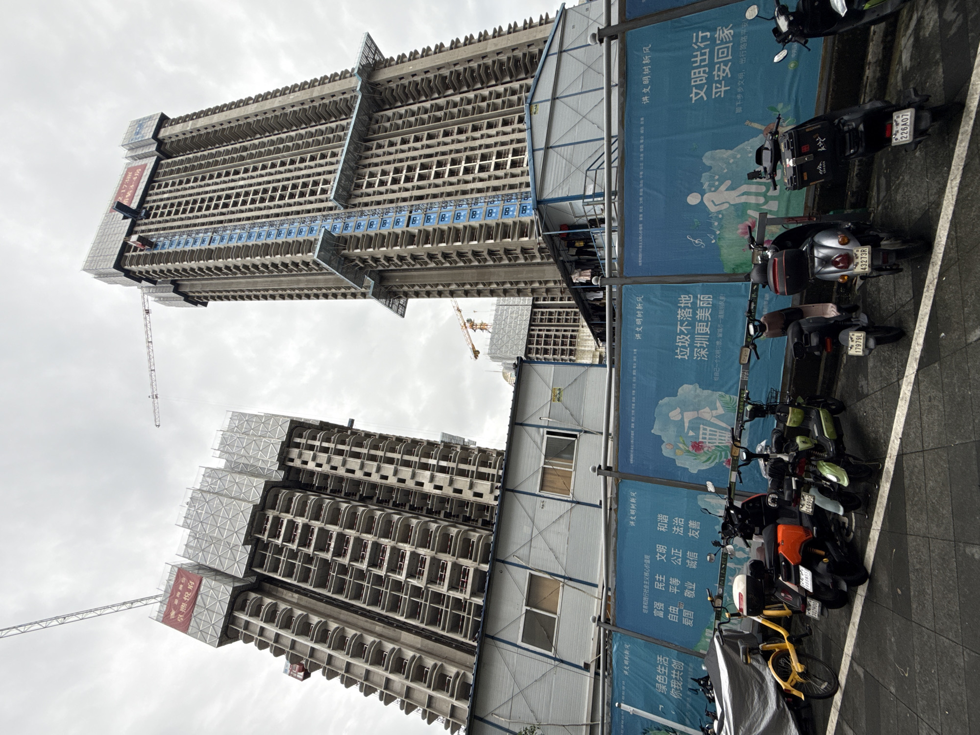 In the city of Futian District, China, an outdoor construction site is depicted under an overcast sky. Two tall, unfinished high-rise buildings, constructed primarily of concrete and metal scaffolding, dominate the scene. Cranes are visible, indicating ongoing construction. In the foreground, a row of parked electric scooters and one bicycle are lined up. Behind the vehicles, a blue banner with Chinese text and graphics, including images of people and recycling, is stretched across a temporary structure. The temporary structure appears to be an office or storage area for the construction site, with windows and laundry hanging outside. The ground is paved with dark tiles.