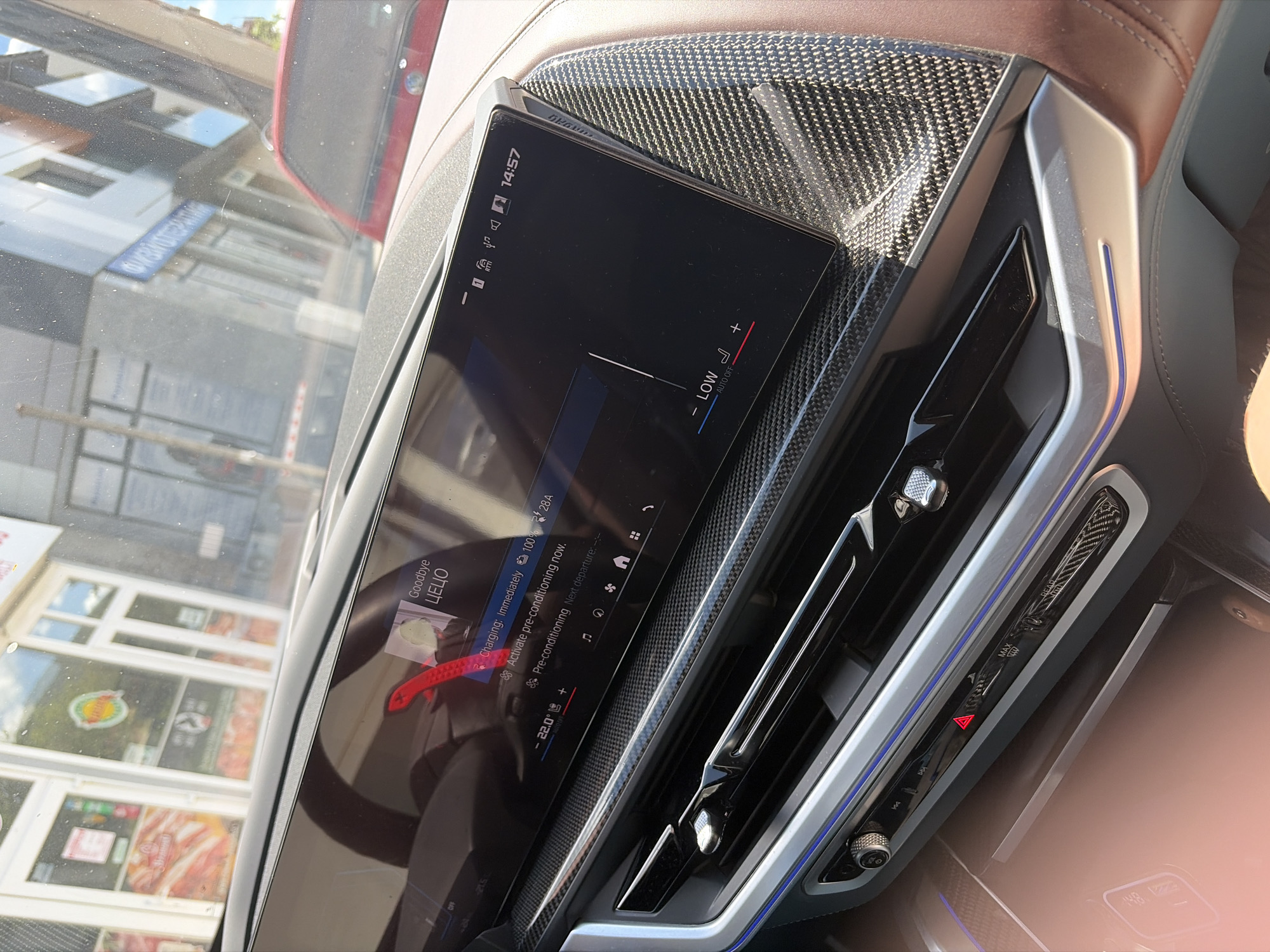 The image depicts the interior of a car, focusing on the dashboard area. A large display screen shows various information, including a song title "Goodbye ЦЕЦО" and details about the car's charging status (100%, 28A). It also indicates the option to activate pre-conditioning and displays the current temperature setting as 22.0 degrees. Below the screen, there are air vents and controls for adjusting the airflow and temperature. The dashboard itself has a carbon fiber pattern. Through the windshield, the exterior reveals a view of a building, likely a shop or restaurant, in what appears to be a busy urban area. The building features large glass windows, and its signage is partially visible. From the available information and clues from the image, the location is Shumen, Bulgaria, given the song title is in Cyrillic.