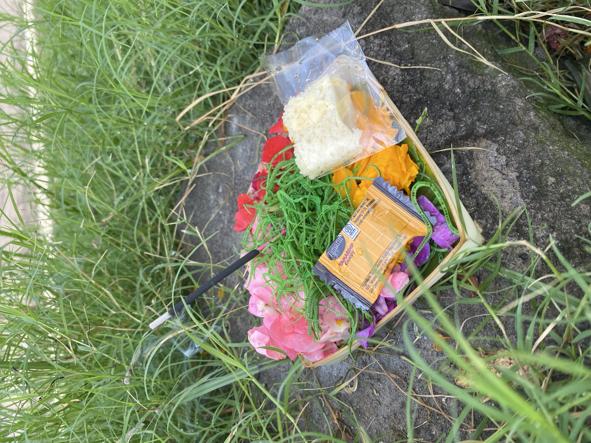The image shows a Balinese offering, a "Canang Sari," placed outdoors on a concrete surface surrounded by grass, likely in Nusa Dua, Indonesia. The offering is in a small, square woven container. Inside, there are several items: assorted flowers (pink, red, and purple), green decorative grass, pieces of what appears to be white cake in a clear plastic bag, an orange sweet (possibly squash or pumpkin), and a packet of "Tamarind Candy." An unlit black incense stick is positioned at the side, partially obscured by the grass. The text on the candy wrapper reads "Tamarin INDAH TIK Tangerang 15135, Indonesia." There are no people visible in the image.