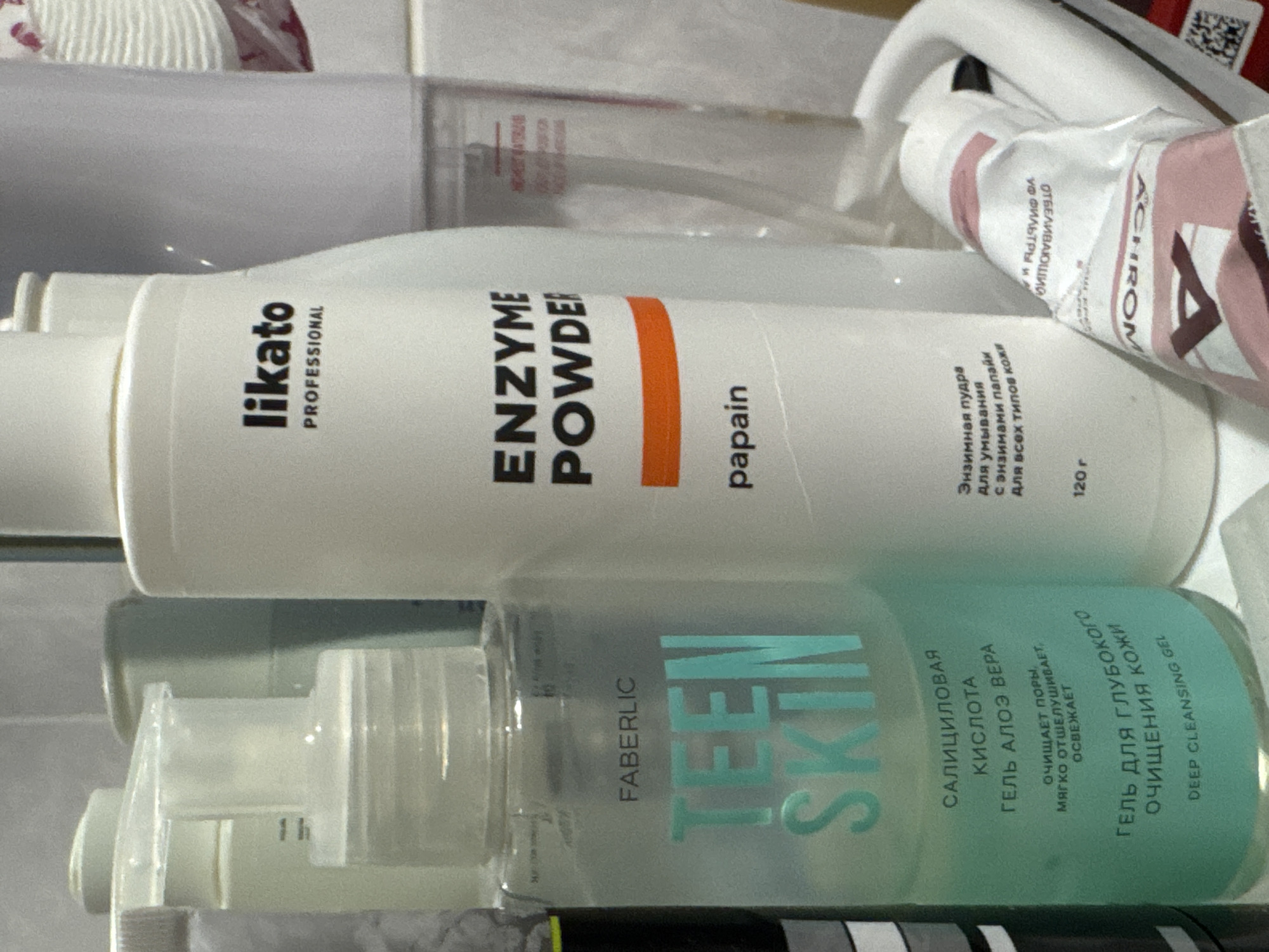 The image shows a close-up of various skincare products arranged on a shelf, likely in a bathroom. In the foreground, a teal-colored bottle labeled "FABERLIC TEEN SKIN" contains a clear liquid. To its right, a white bottle with black lettering reads "likato PROFESSIONAL" and "ENZYME POWDER papain". Further to the right, a white tube with pink and red accents is partially visible, with text that appears to be in a foreign language. The background consists of muted, neutral colors, suggesting a domestic setting. The arrangement of the products and the lighting indicate a still-life composition. There is no indication of the specific location within Яромінскі сельскі Савет, Belarus, or any people or actions.