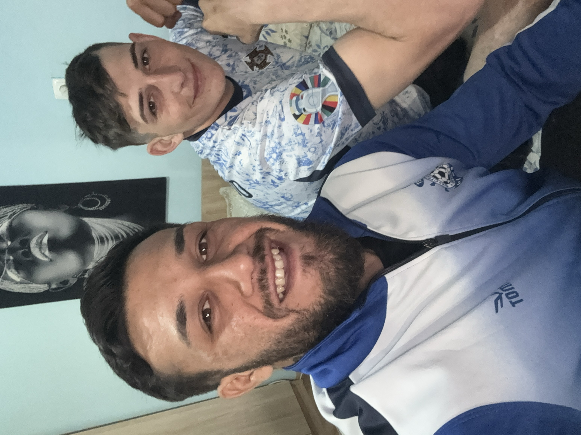 Two men are posing for a selfie. One man is wearing a white and blue sports jacket. The other man is wearing a white and blue soccer jersey with the Portugal crest and the logo for the UEFA European Championship. The location cannot be determined from the image.