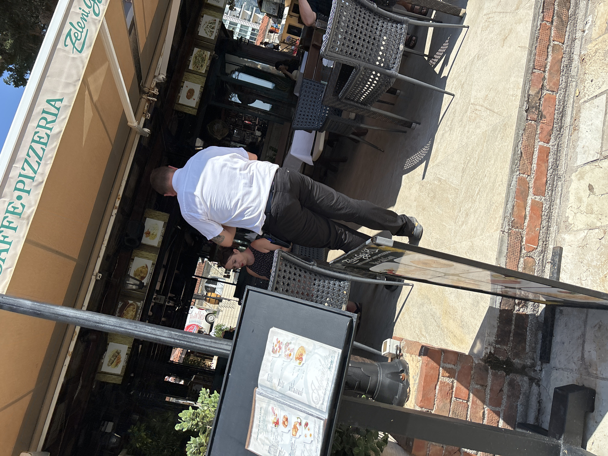 This media file shows an outdoor seating area of a cafe or restaurant, likely in Budva, Montenegro. The scene is oriented sideways, so a person in a white shirt and black pants is the primary subject, viewed from behind as they lean over a table. They appear to be a waiter or staff member, possibly presenting a menu or attending to a customer.
Another person, a woman wearing a black and white polka dot top, is seated at a table, looking down and to the side. The tables are made of dark woven material, with metal chairs. A large awning stretches over the seating area, providing shade.
In the background, there are glimpses of buildings, suggesting an urban or town setting. The ground is paved with light-colored tiles and some brickwork. A menu is open on a black table in the foreground, displaying pictures of food items.
The lighting suggests it is daytime, with strong sunlight casting shadows. Visible text on the awning reads "ZELENI G..." and "CAFE PIZZERIA". The menu also has text, but it is not clearly legible. The overall impression is a casual outdoor dining or service environment.
