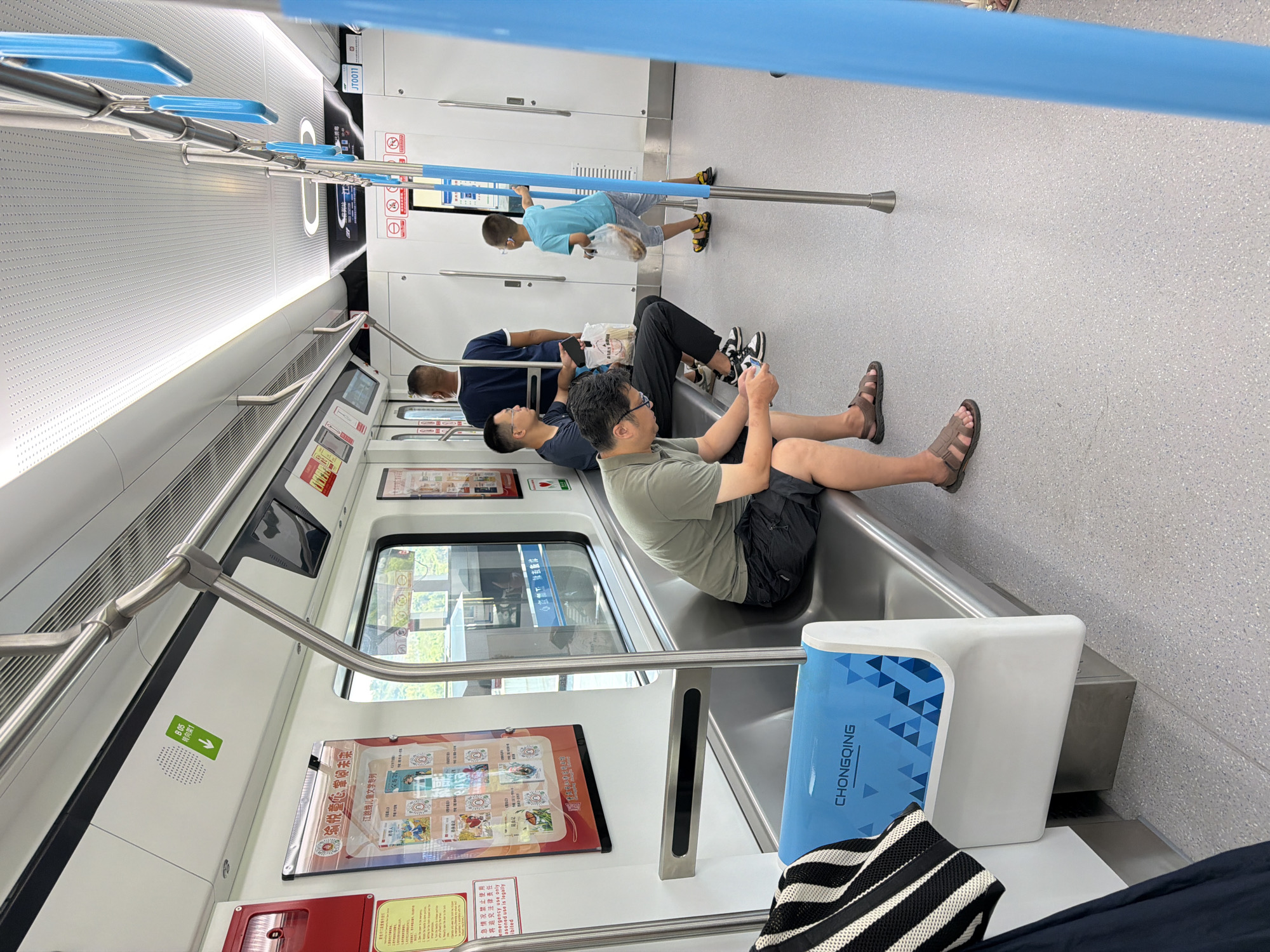 The image depicts the interior of a subway train car in Jiangjin, China. Several people are seated on the bench-style seats, which are metallic and run along the length of the car. One man in the foreground, wearing a green t-shirt and black shorts, is seated and looking down at his phone, wearing sandals. To his left, another man in a dark blue shirt and black pants is also seated, holding a phone and a plastic bag. Further down the car, a young boy in a blue t-shirt and grey shorts stands near the doors, holding a plastic bag. A man in a dark blue t-shirt stands behind him. The train car interior is brightly lit with overhead fluorescent lights and features metallic grab bars and blue hand straps for passengers. Posters are displayed on the walls, and the floor is a light gray speckled material. The overall environment is clean and modern.