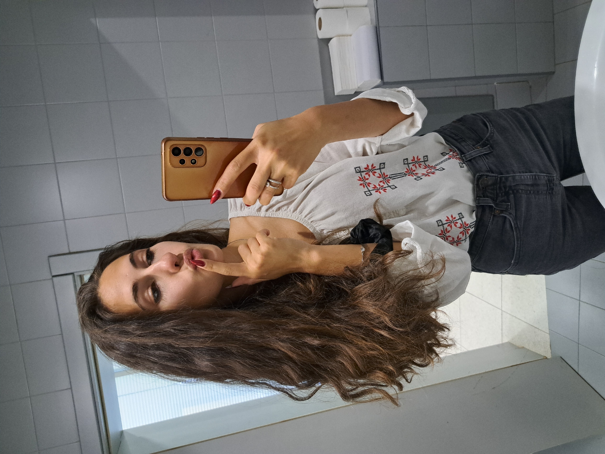 A young woman is taking a selfie in what appears to be a bathroom. The bathroom has a light gray tiled wall and a white sink. She is holding a bronze-colored smartphone with multiple camera lenses. She is wearing a white blouse with red and black embroidery and gray jeans. She is touching her lips with her index finger. Her hair is long and brown. It's impossible to determine the specific location in Sofia, Bulgaria, without further information or context within the image. The bathroom provides no direct indication of its location.