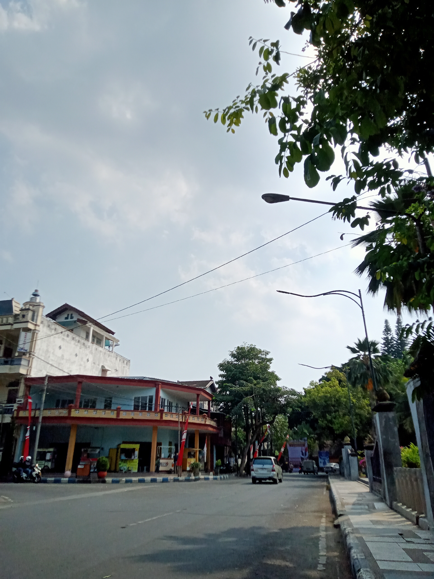 The image depicts a street scene in Tulungagung, Indonesia, on a clear day with scattered clouds. On the left, a multi-story building with white and red accents stands prominently, featuring balconies and several windows. Two red flags are visible on poles in front of this building. Below the balconies, there are several small food stalls or kiosks, one yellow and two red. To the left of the building, two motorcycles are parked, and one person is visible on a motorcycle. A silver sedan is driving away from the camera on the road in the center of the image. On the right side of the road, a sidewalk with patterned paving stones runs alongside the street. Further down the street, several trees and palm trees create a lush green backdrop, with a tall concrete wall and pillars on the far right. Two streetlights are visible in the sky, one with a curved pole and the other straight. Branches of a leafy tree overhang the top right of the frame, with sunlight filtering through the leaves.