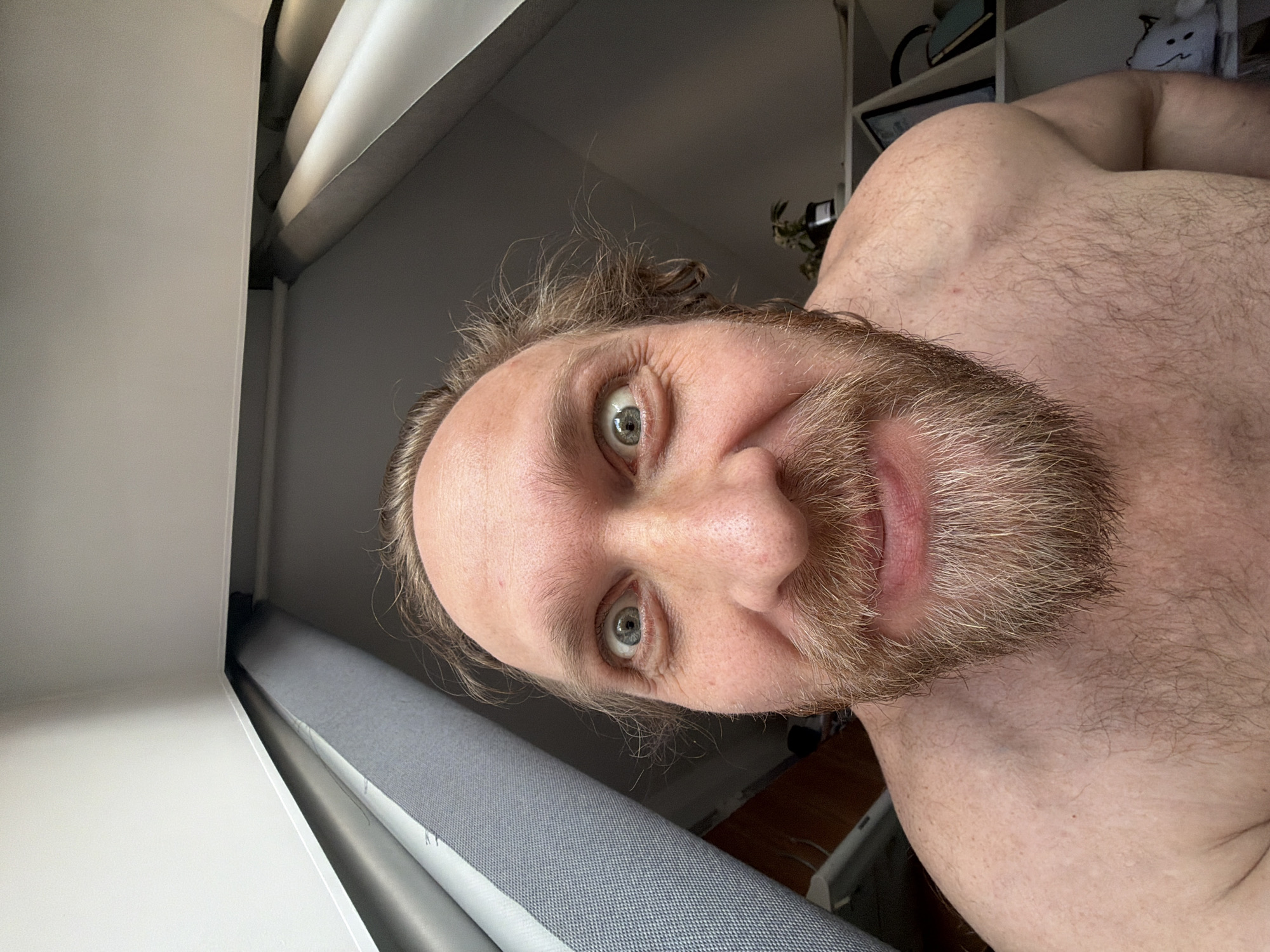 A man with a beard and light eyes is visible, appearing to be in an indoor environment near a window. The image includes portions of his bare chest and shoulder. The surroundings are indistinct, but show elements such as white walls, what might be window blinds, and shelves in the background. There are no indications of Saint Petersburg, Russia, or any specific location within it.