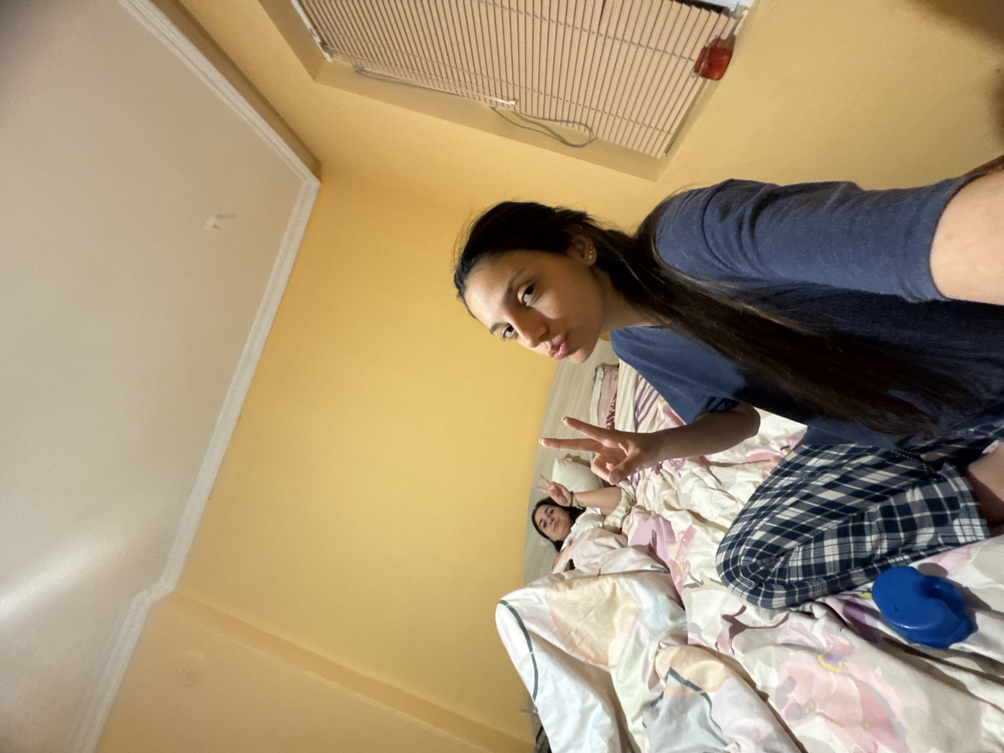 This indoor photograph captures a casual and playful moment featuring two young women, likely in a bedroom in Drenovets, Bulgaria. The image is taken from a low angle, giving it a selfie-like perspective.
In the foreground, a young woman with long dark hair, wearing a dark blue t-shirt and blue plaid pajama bottoms, is partially visible. She is making a peace sign with her right hand, her fingers prominently displayed, and has a slight pout on her face, looking towards the camera. Behind her, another young woman is lying in a bed with floral-patterned bedding, also making a peace sign with her hand. She appears to be smiling or looking relaxed.
The room features light yellow walls and a white ceiling, with a white decorative trim where the wall meets the ceiling. A window covered by horizontal blinds or shutters is visible in the upper part of the frame. A small, rounded blue object, possibly a hair accessory or a small container, rests on the bed near the foreground subject's leg.
The overall mood is relaxed and informal, suggesting a friendly interaction between the two individuals. The lighting indicates an indoor setting, potentially during the evening or night. No discernible text is visible in the image.