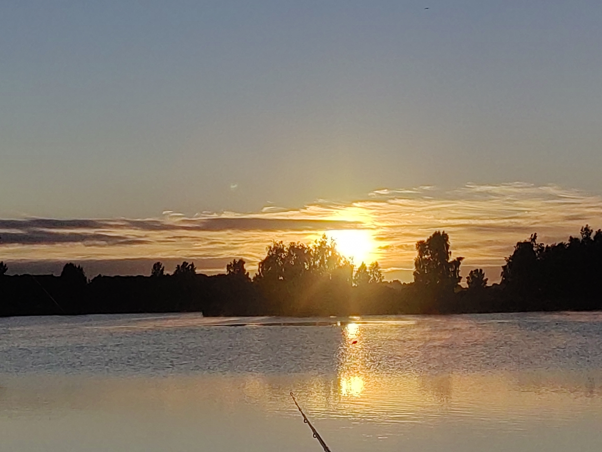 The image captures a sunset over a body of water, likely a lake or river, in the vicinity of Усяжскі сельскі Савет, Belarus. The sky transitions from a pale blue at the top to hues of orange and yellow near the horizon, with scattered clouds catching the last rays of sunlight. A silhouetted treeline, with distinct tree shapes, forms the background against the vibrant sky. The water's surface reflects the warm colors of the sunset, creating a shimmering effect. In the foreground, a fishing rod is visible, partially submerged in the water, suggesting a fishing activity is taking place. A small red float is also present on the water's surface, further indicating fishing. The overall scene depicts a tranquil, natural environment during the twilight hours.