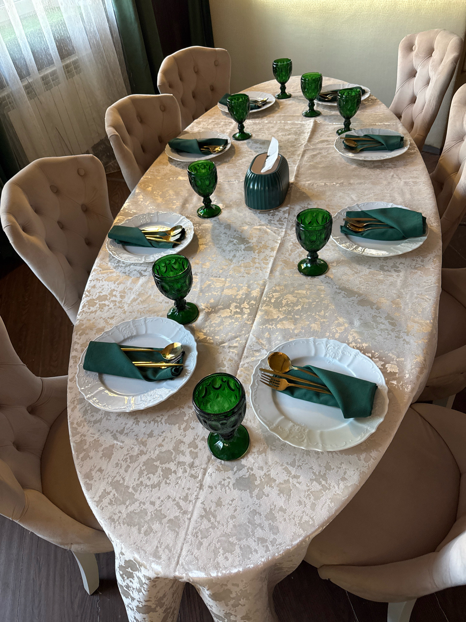 An overhead, eye-level shot captures a dining table set for multiple people in what appears to be a restaurant in Torzhok, Russia. The table is covered with a cream-colored, patterned tablecloth. On the table, there are eight place settings, each featuring a white plate with an embossed floral design. A dark green cloth napkin is folded on each plate, with gold-colored cutlery (a fork and a spoon) placed on top of the napkin. There are also eight emerald green stemmed glasses arranged around the table. One place setting in the center of the table has a green tissue holder with white tissues. The background shows the backs of several upholstered dining chairs in a neutral beige color. To the left of the table, sheer white curtains hang over a window, revealing the faint outlines of blinds and a radiator. The flooring is a dark wood. The lighting appears to be natural light from the window and overhead artificial light, creating a warm ambiance.