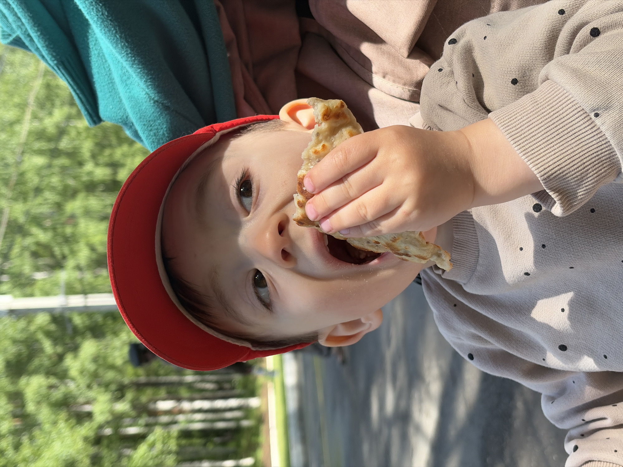A toddler wearing a red baseball cap and a light brown, polka-dotted sweatshirt is eating a piece of flatbread. The child is outside, with a blurred background of green trees and a gray paved area. The image appears to have been taken in an urban or suburban environment. There is no specific evidence to confirm this is Izhevsk, Russia.
