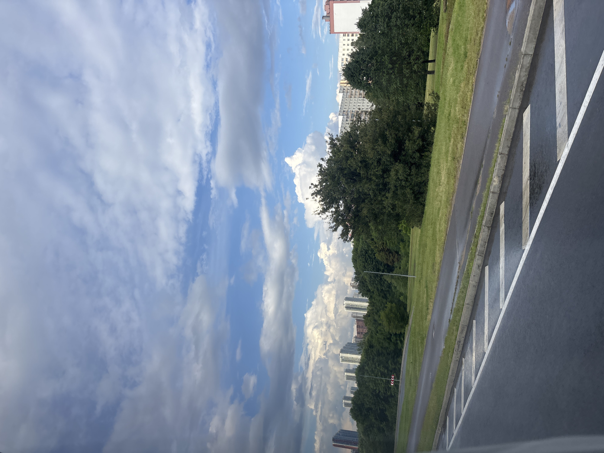 The image depicts a cityscape with residential buildings visible in the background, partially obscured by lush green trees. The foreground features a wet asphalt road with distinct white lane markings, suggesting recent rain. The sky is a mix of blue and dramatic grey clouds, indicating potentially changing weather conditions. The scene appears to be an urban or suburban environment within the Сеніцкі сельскі Савет, Belarus, characterized by a blend of natural greenery and modern architecture.