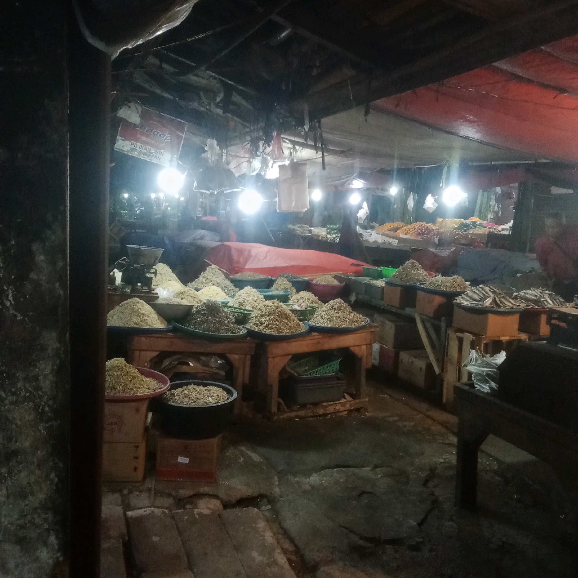 This image depicts an outdoor market stall at night, likely in Karawang, Indonesia. The stall is covered by a red tarpaulin, supported by wooden beams. The ground appears to be made of concrete or packed earth, showing signs of wear and possibly moisture.
The primary focus of the stall is the display of dried goods, predominantly small fish and what appear to be grains or seeds, arranged in large platters and bowls. Several wooden crates are visible beneath the platters, some containing more of the dried goods. A vintage scale sits on a table behind one of the piles of goods.
Lighting comes from several bare bulbs, casting a harsh, bright glow on the products and illuminating the scene. The overall atmosphere is dim and somewhat cluttered, characteristic of a local market at closing or preparing for opening hours.
In the background, glimpses of other stalls and the shapes of people can be seen, suggesting a larger market area. One individual is partially visible on the right side of the frame, wearing a dark top. The overall impression is a functional, working market environment.