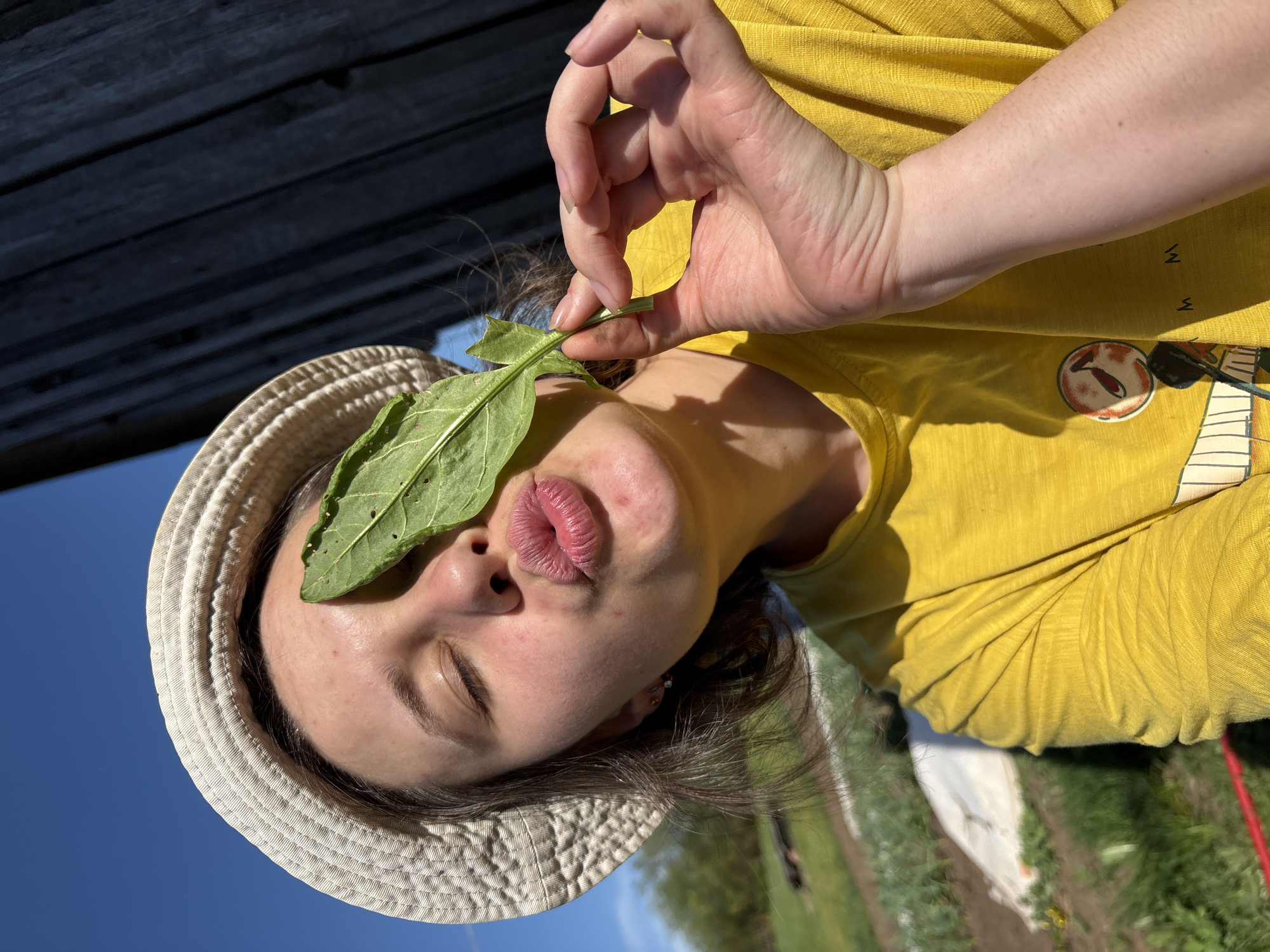 A woman wearing a white sun hat and a yellow t-shirt is holding a green leaf over her closed right eye, pursing her lips in a kissing gesture. Her hand is making an "OK" sign. The photo appears to have been taken outdoors, with a partially visible wooden structure above and a blurred background of greenery and blue sky, suggesting a garden or park setting. The location is not discernable from this image.