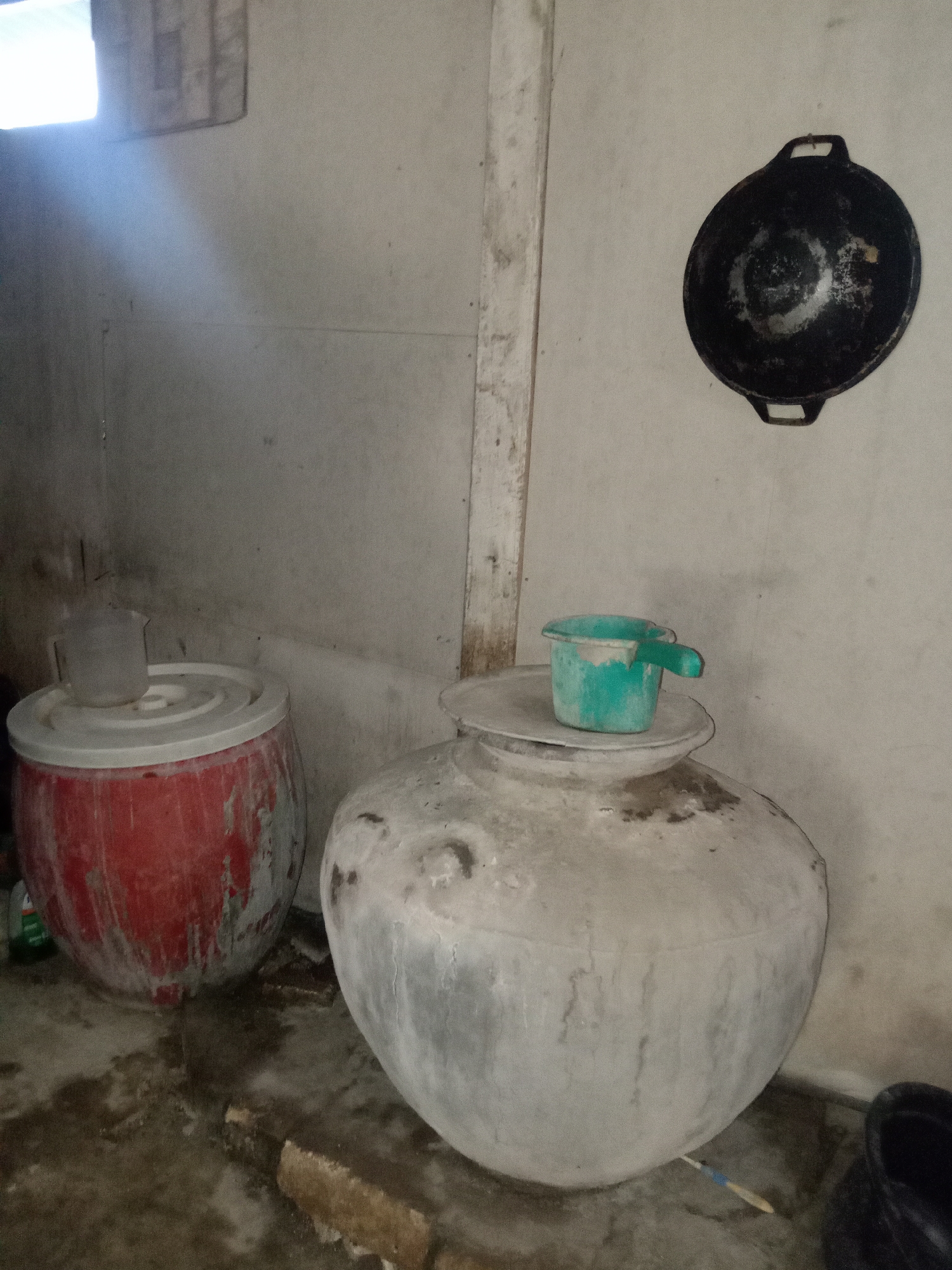 A ground-level, medium shot captures an indoor scene with several large containers and cooking implements. In the foreground, two large, round, light-colored concrete urns sit on a damp, concrete floor. The urn on the right is the largest, with a light gray, weathered surface. Perched on top of it is a small, teal-colored plastic scoop with a handle. To the left of this large urn is another large urn, red in color and also weathered. On top of this red urn rests a clear, plastic pitcher. Behind these urns, a light-colored wall dominates the background, with a wooden beam running vertically down the center. On the upper right portion of the wall, a dark, circular cooking pan with two handles is mounted. A bright light source is visible in the upper left corner, casting a diagonal ray of light across the wall. The floor appears wet and stained, suggesting a functional, possibly utilitarian space. The scene is consistent with a domestic storage area or a kitchen in a humid climate. The environment suggests a practical, everyday setting, possibly in Stiaji, Indonesia, where such large water storage vessels are common.