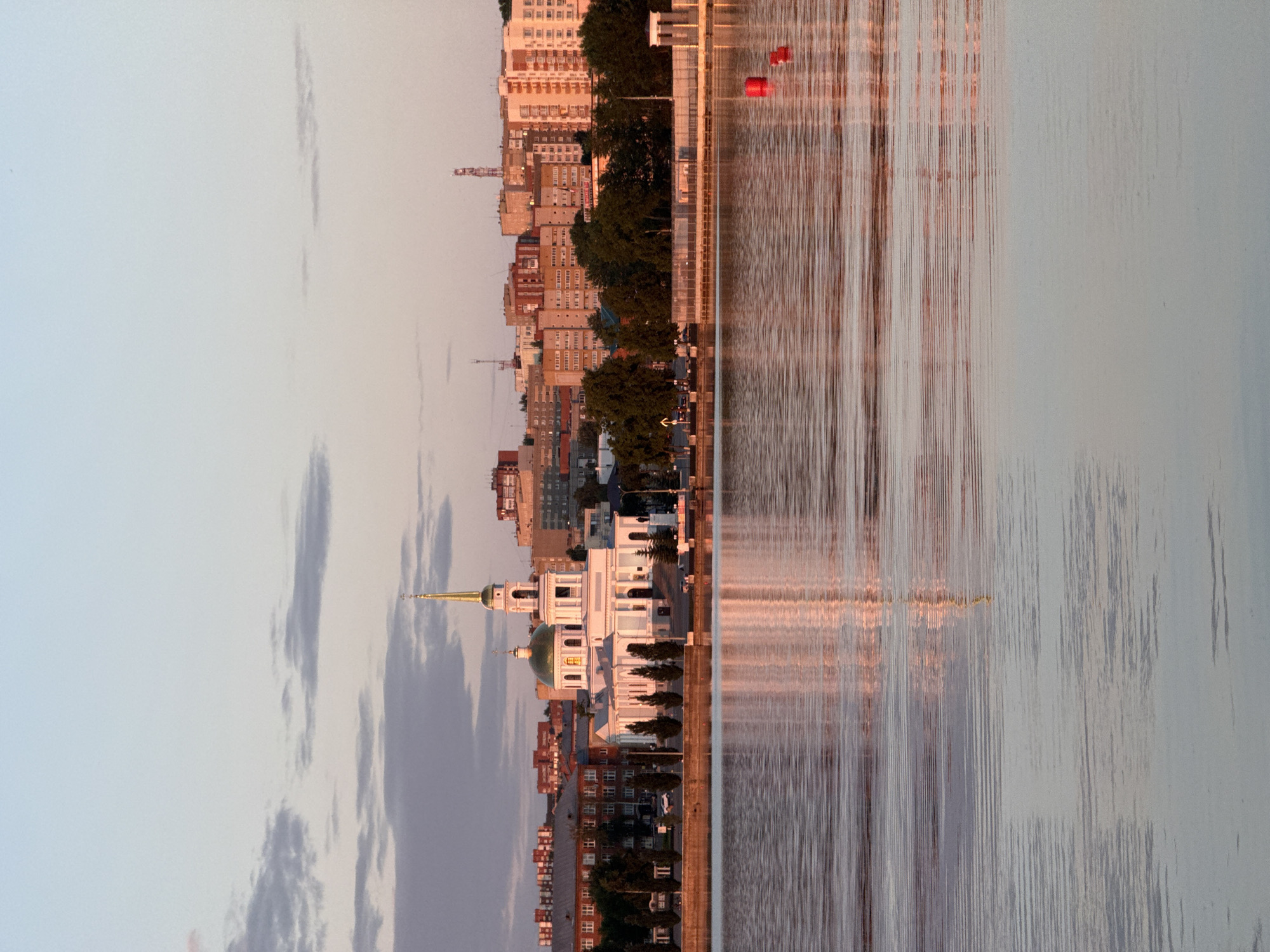 The image depicts a serene urban landscape at either sunrise or sunset, looking across the Votkinsk Pond towards the city of Votkinsk, Russia. The central feature is the white-painted St. Elijah Church (Il'inskaya Tserkov), prominently situated on the northern bank of the pond. The church features a large green dome, a tall white bell tower, and a slender golden spire topped with an Orthodox cross, all illuminated by warm, low-angle light.
To the left of the church, a row of multi-story buildings, including brick and pastel-colored residential structures, extends along the shoreline. To the right of the church, the embankment features a promenade lined with slender trees, behind which further urban development, including more multi-story buildings, is visible. Several communication towers are discernible on the distant skyline.
The foreground is dominated by the calm, rippled surface of the Votkinsk Pond, which beautifully reflects the warm hues of the sky and the illuminated structures on the bank. The reflections of the church's spire and the surrounding buildings are visible in the water. Two distinct red cylindrical buoys are floating on the right side of the pond. The sky transitions from a pale blue at the zenith to soft pink and orange tones near the horizon, with scattered, thin grey clouds. No individuals are clearly visible in the scene, and no specific actions are discernible. The overall impression is one of tranquility and urban harmony with nature.