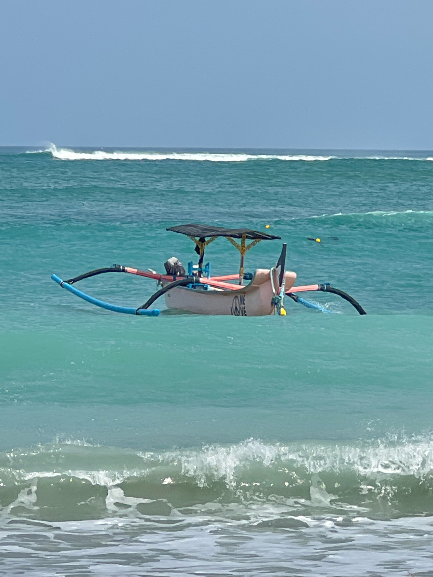 A pink and white outrigger canoe floats on turquoise water. The canoe has a black canopy, a propeller, and two outrigger floats. The water is calm with gentle waves. In the distance, a larger wave is breaking. The sky is clear and blue. This scene is likely Kuta Beach, Bali, Indonesia, known for its surf and local fishing boats.