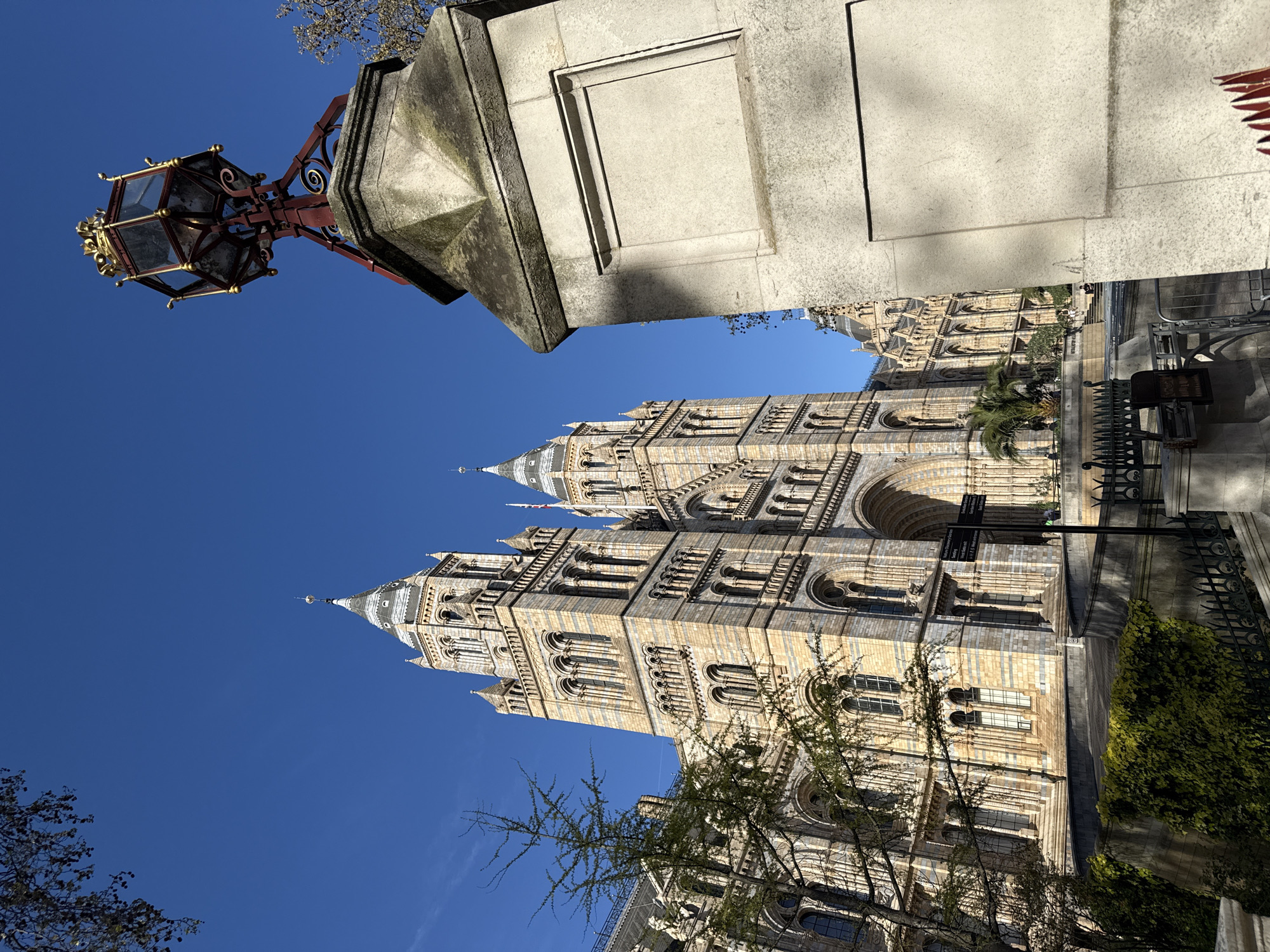 The image shows a building under a bright blue sky in London, UK. The architecture is ornate, with towers and numerous windows. A light post with an ornate lantern sits on a stone base in the foreground. Part of a bushy green tree is along the building's side. A fence is in the background.
